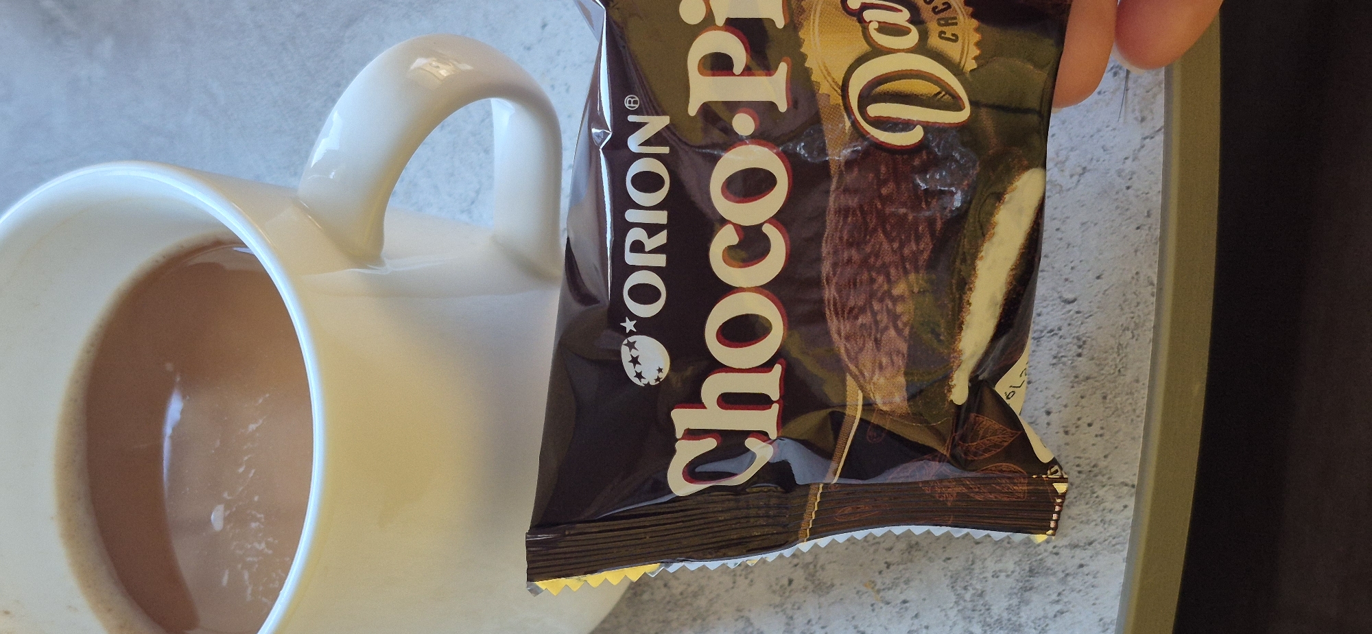 A partially full white mug containing a light-brown liquid sits adjacent to a brown Orion Choco-Pie Dark package. The mug's handle is visible and extends outward. The Choco-Pie package is held in someone's fingers. The scene takes place in an unidentified location in the city of Unknown, Unknown.