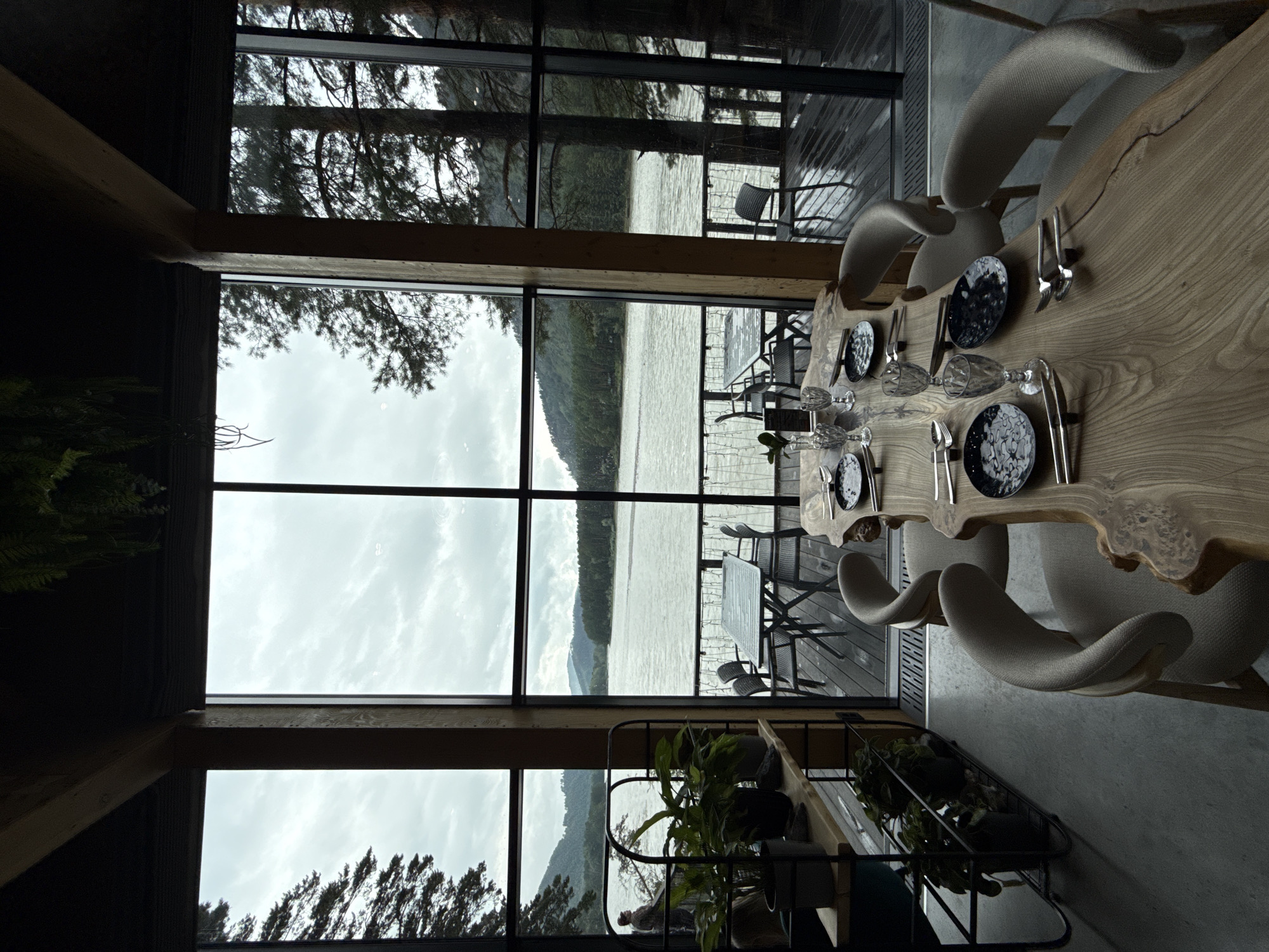A dining area is situated indoors with a panoramic view of a wide river and forested hills under a cloudy sky. The room features large windows that offer an unobstructed vista. In the foreground, a long, live-edge wooden table is set for a meal with plates, glasses, and cutlery. Several upholstered chairs surround the table. To the left, a shelving unit holds potted plants. Outside the windows, a wooden deck is visible, furnished with tables and chairs. A person stands on the deck looking out at the river. The scene suggests a serene, natural setting, possibly a restaurant or lodge. The location is identified as Nizhnekayancha, Russia.