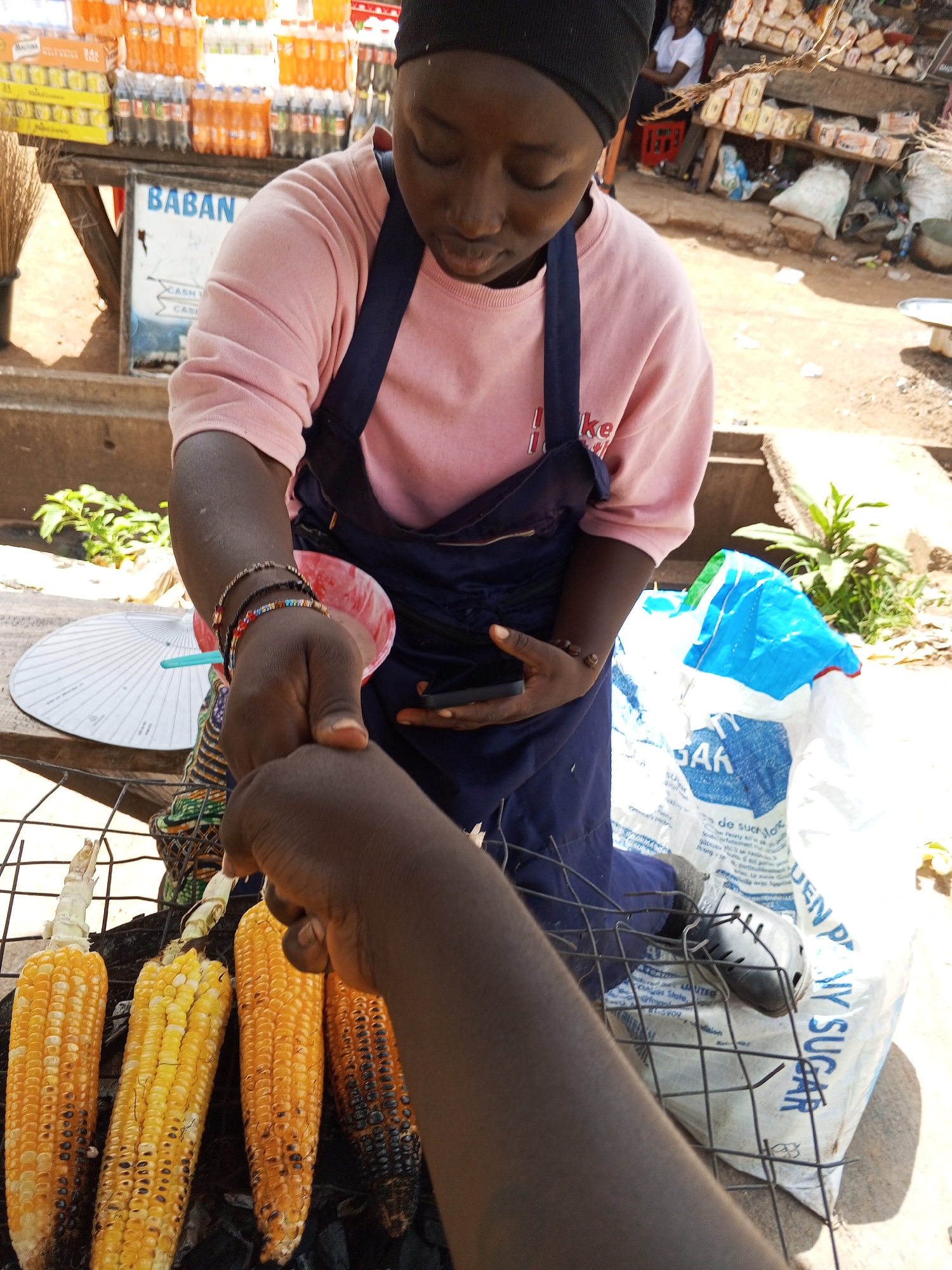 In the city of Bukuru, Nigeria, a young Black individual wearing a pink sweatshirt, a dark blue apron, and a black hijab is selling grilled corn. The individual is holding a piece of corn, seemingly in the process of handing it to another person whose arm and hand are visible in the foreground. Several ears of grilled corn are arranged on a wire rack over coals. In the background, there is a stall with various bottles of beverages and packaged goods. A white fan is also visible. The overall environment appears to be an outdoor market or street vendor setting.