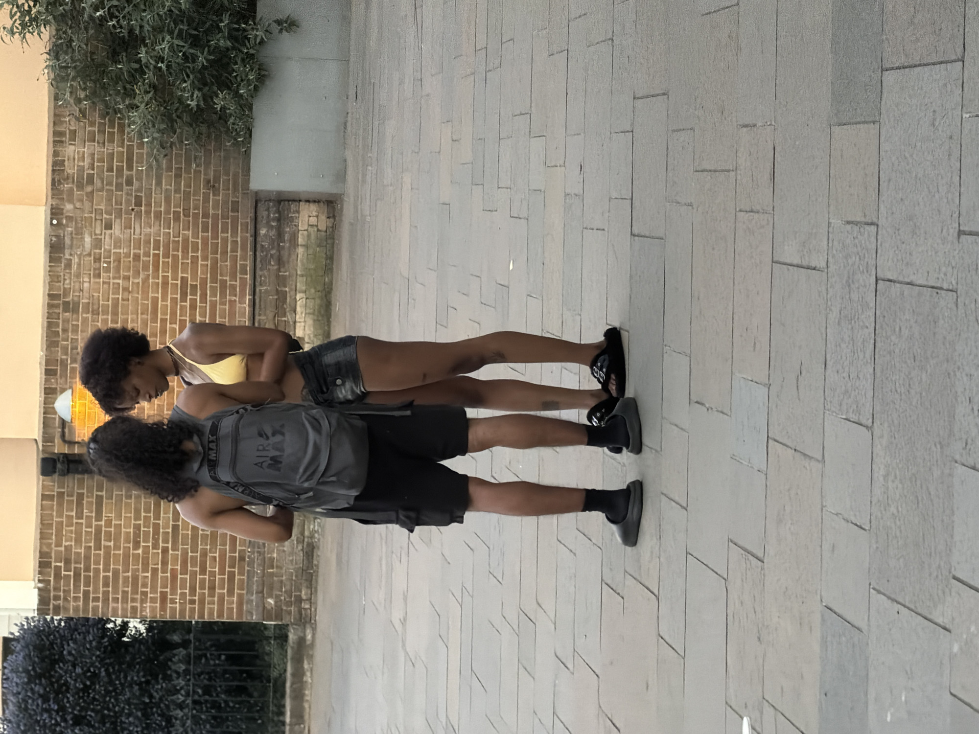 Two young adults stand on a sidewalk paved with large, light-colored rectangular stones. The adult in front wears a yellow top and jean shorts. Behind the other, the second adult wears a gray backpack labeled "Air Max," a dark shirt and shorts, and dark socks with clogs. Both stand against a background of a brick wall and greenery. The location could be London, United Kingdom.
