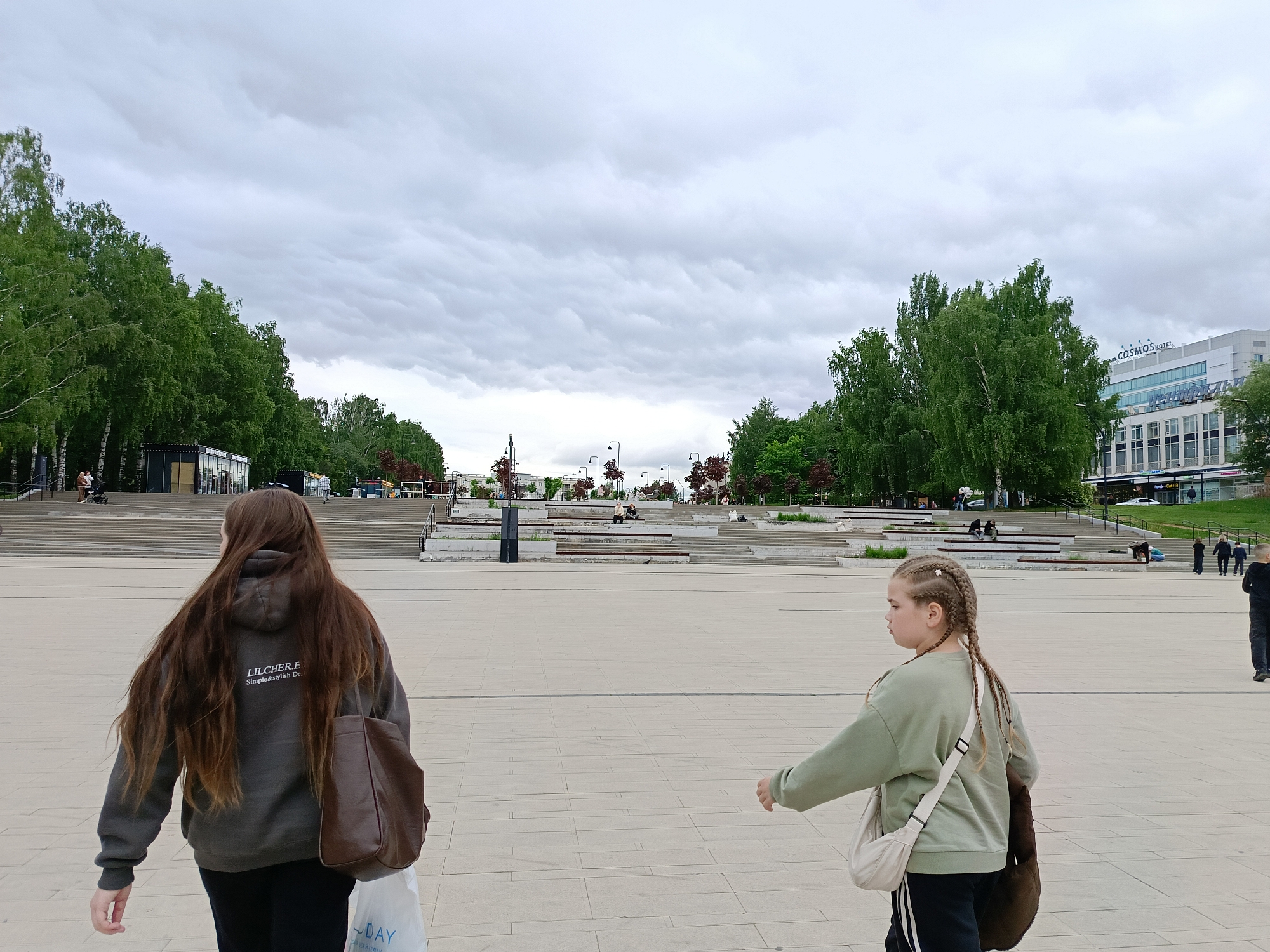 This image depicts an expansive urban plaza in Izhevsk, Russia, situated near the Central Square, with the Cosmos Hotel prominently visible in the background. The scene is observed on an overcast day, characterized by a sky filled with grey clouds.
In the immediate foreground, two individuals are walking across a vast, light-colored paved surface composed of square tiles. On the left, a person with long, straight brown hair, seen from behind, wears a dark grey hooded sweatshirt. The back of the sweatshirt features white text that reads "LIL.CHER.E Simple&stylish Des." This individual carries a brown leather shoulder bag on their right side and a white plastic shopping bag with blue text in their left hand. On the right, a young person with light brown hair styled in multiple tight braids, some adorned with white decorative elements, walks with their head turned slightly to the left. They are dressed in a light green long-sleeved sweatshirt and dark pants with a vertical white stripe down the side, carrying a light-colored crossbody bag and a dark, possibly brown, soft shoulder bag.
The midground features a wide series of concrete steps and terraced platforms, with some sections equipped with dark metal railings, ascending towards the distant background. Scattered across these levels are several black, modern streetlights. Multiple other individuals are visible in the midground and background, either walking up or down the stairs, or seated on the platforms.
The background is defined by dense lines of green trees, including several birch trees with their characteristic white bark, flanking the plaza. To the far right, a modern, multi-story building with a light-colored facade displays the blue illuminated sign "COSMOS HOTEL" and additional text. On the left side of the plaza, near the tree line, a few small, dark kiosk-like structures are visible. The environment conveys a sense of a large, contemporary public space.