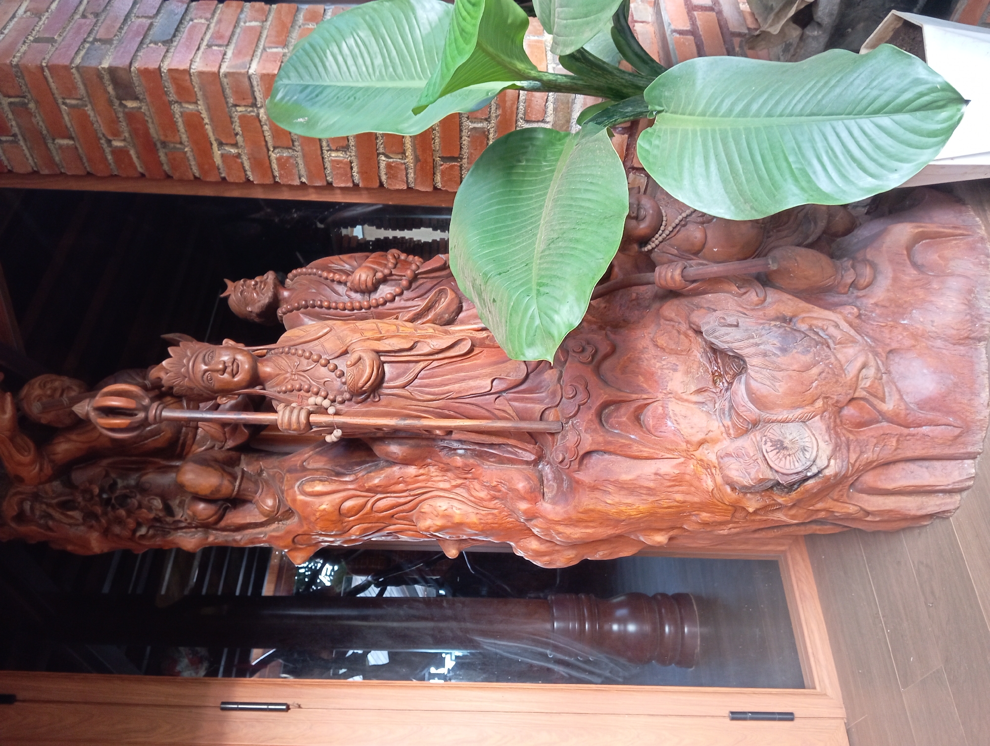 In the city of Chư Sê, Vietnam, a wooden sculpture stands prominently near a brick wall. The sculpture, carved from reddish-brown wood, depicts several figures. The central figure is a tall, robed person wearing a crown and holding a staff. To their right, another figure with a beard and wearing a cap is visible. Below these figures, a smaller, smiling figure is partially obscured. The sculpture is intricately detailed, with flowing robes and beaded necklaces adorning the figures. The base of the sculpture is rough and textured, resembling rock formations. Adjacent to the sculpture, a lush green plant with large leaves in a white pot adds a touch of nature to the scene. A wooden door with hinges is on the left, and a reflective surface suggests a window or a glass door, revealing glimpses of the interior, including wooden columns and possibly other decorative elements. The ground is a wooden floor. The overall setting appears to be an outdoor or semi-outdoor area, possibly part of a cultural or religious site given the nature of the sculpture.