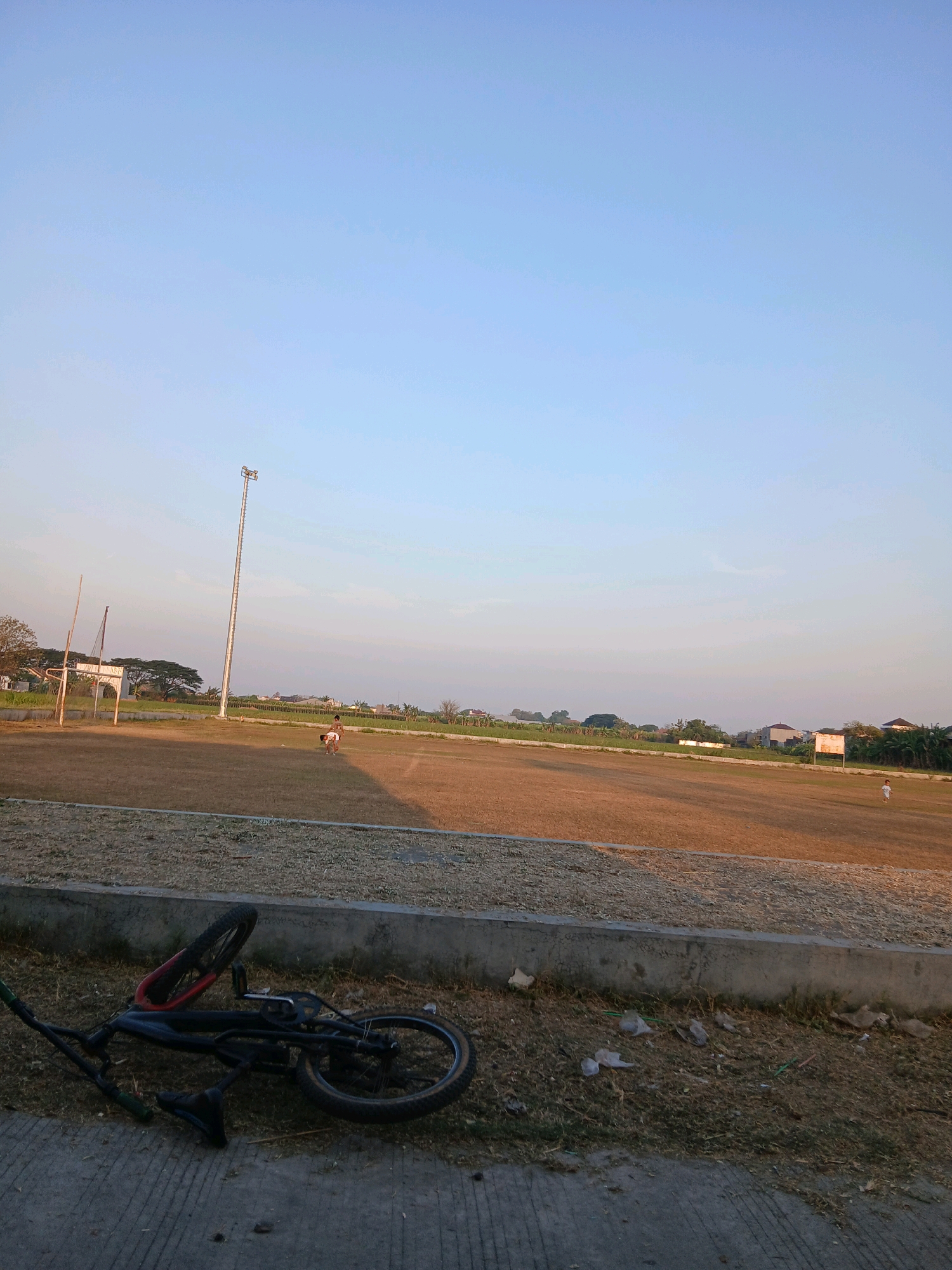 An outdoor, horizontal shot shows a scene at a sports field with a clear, pale blue sky. In the foreground, a black BMX bicycle lies on its side on a concrete path, with its handlebars, seat, and wheels clearly visible. The bicycle is near a strip of dry grass and weeds, which borders a concrete retaining wall. Beyond the wall is a wide, dry, brown grass field, likely a sports pitch. A tall light pole stands prominently on the field, with lights at its apex. In the middle ground, two young individuals, one appearing to be an adult and the other a child, are visible on the field. The adult is in a crouched or bent-over position, possibly interacting with the child, who is standing. Further in the distance, on the right side of the field, another small figure, likely a child, is standing. The background shows a line of trees and residential buildings under the expansive sky. The scene appears to be a public sports facility located in the vicinity of Jepangpakis, Indonesia, likely during late afternoon or early evening due to the soft, warm lighting.