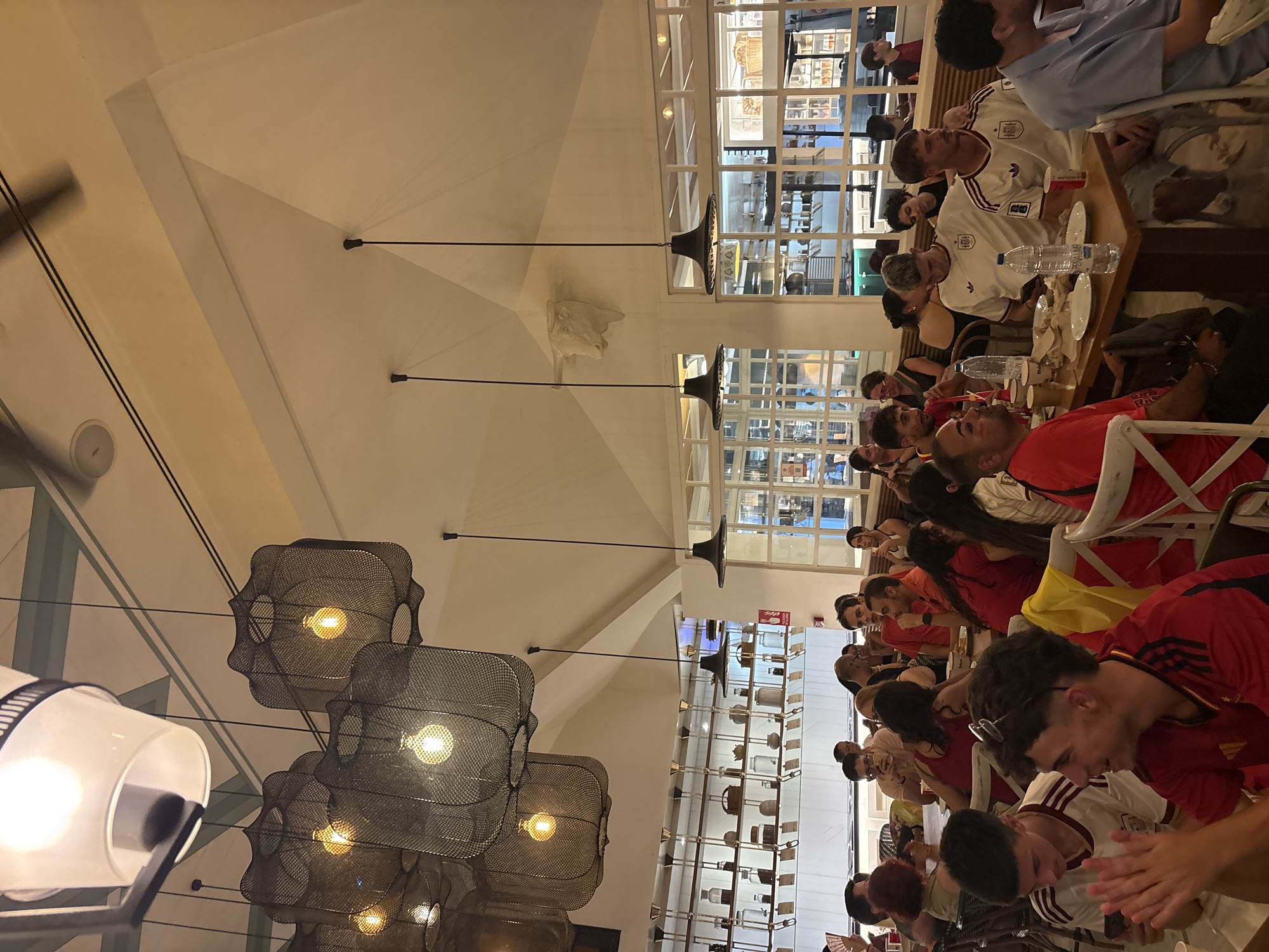 The image depicts an indoor scene within a restaurant, likely in the city of Valencia, Spain, given the context provided by the people wearing what appear to be Spanish national team jerseys. The restaurant has a high ceiling with decorative lighting fixtures, including large, dark, mesh-like pendant lamps and a more traditional white shade lamp. A decorative white bull's head sculpture is mounted high on a sloped wall. The dining area is populated by a diverse group of people, many of whom are seated at tables and appear to be engaged in conversation or watching something. Some individuals are wearing red and white jerseys with crests, suggesting they are fans of a sports team. On the tables, there are water bottles, plates, cups, and what seem to be food items. The background reveals shelves lined with various containers and bottles, indicating a serving area or display. The overall atmosphere appears to be social and possibly celebratory.
