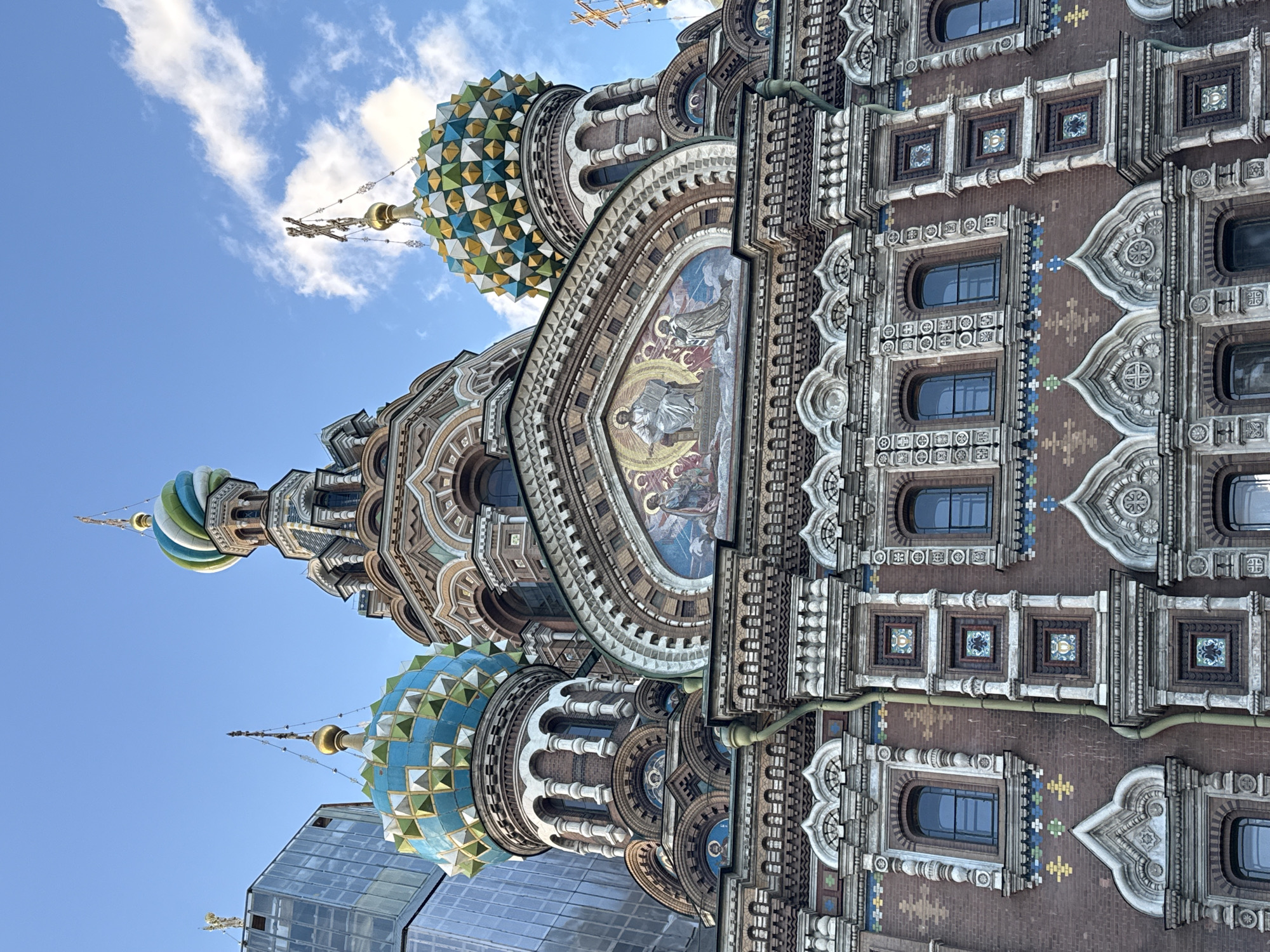 The image shows a low-angle exterior view of the Church of the Savior on Spilled Blood, a Russian Orthodox church located in Saint Petersburg, Russia. The church is characterized by its ornate architecture, featuring multiple onion domes in vibrant colors and intricate mosaic details. The main facade is constructed of red brick and adorned with decorative stonework, arched windows, and various ornamental elements. In the background, a modern glass building with a reflective surface rises, creating a stark contrast with the historic church. The sky above is blue with scattered white clouds. There are no people visible in the image.