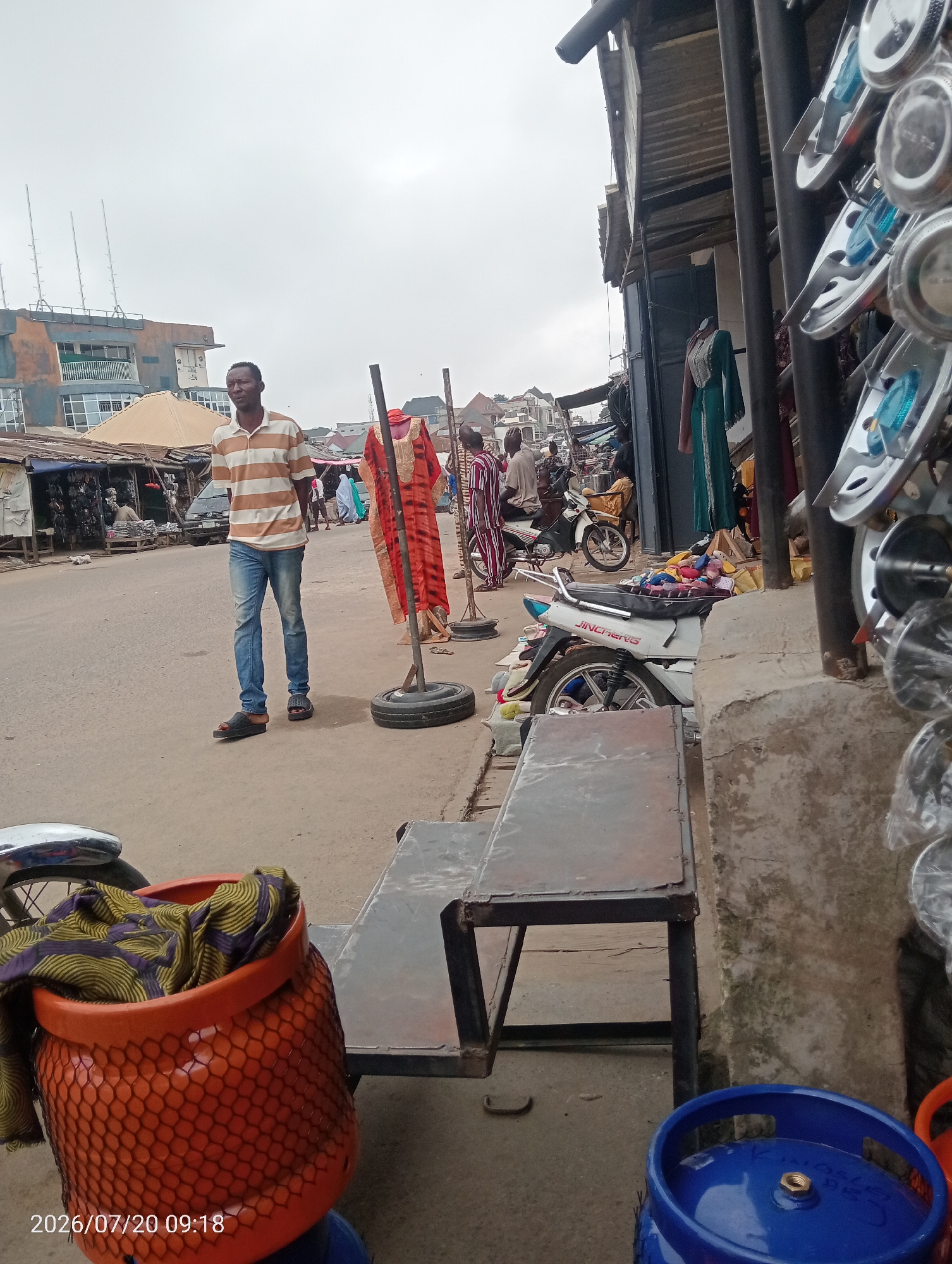 The image depicts a street scene in Suleja, Nigeria, with a focus on a bustling market environment. A man wearing a striped polo shirt and blue jeans stands in the foreground, looking towards the viewer. Behind him, various market stalls are set up along the roadside, displaying clothing and other goods. Several motorcycles are parked on the street, indicating a common mode of transportation. The stalls are constructed from makeshift materials, and the overall atmosphere suggests a vibrant, albeit informal, commercial area. In the background, buildings with corrugated iron roofs and a more substantial structure with large windows can be seen. The sky is overcast, contributing to a muted lighting condition. The presence of a blue gas cylinder and an orange container with fabric in the immediate foreground suggests ongoing daily life and commerce in the area. The timestamp "2026/07/20 09:18" is visible in the bottom left corner.