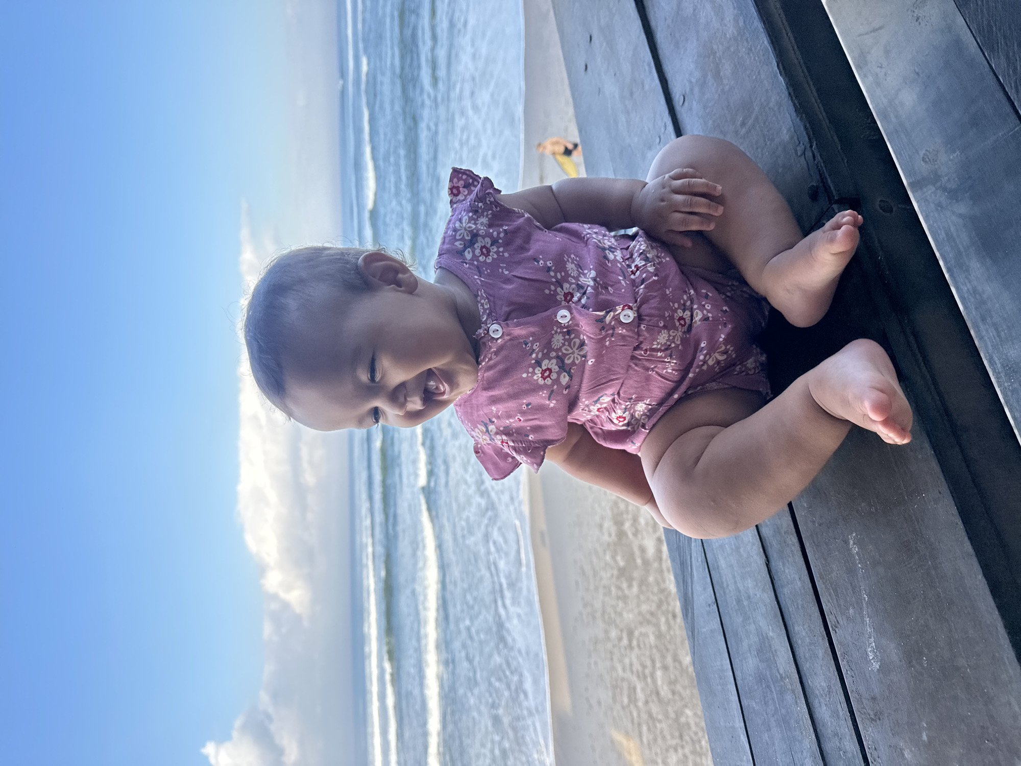 An infant wearing a pink floral romper is sitting cross-legged on a wooden surface. The infant is facing to the right of the frame and is laughing. The background shows a beach scene with the ocean and a cloudy sky. The location is the city of North Kuta, Indonesia.