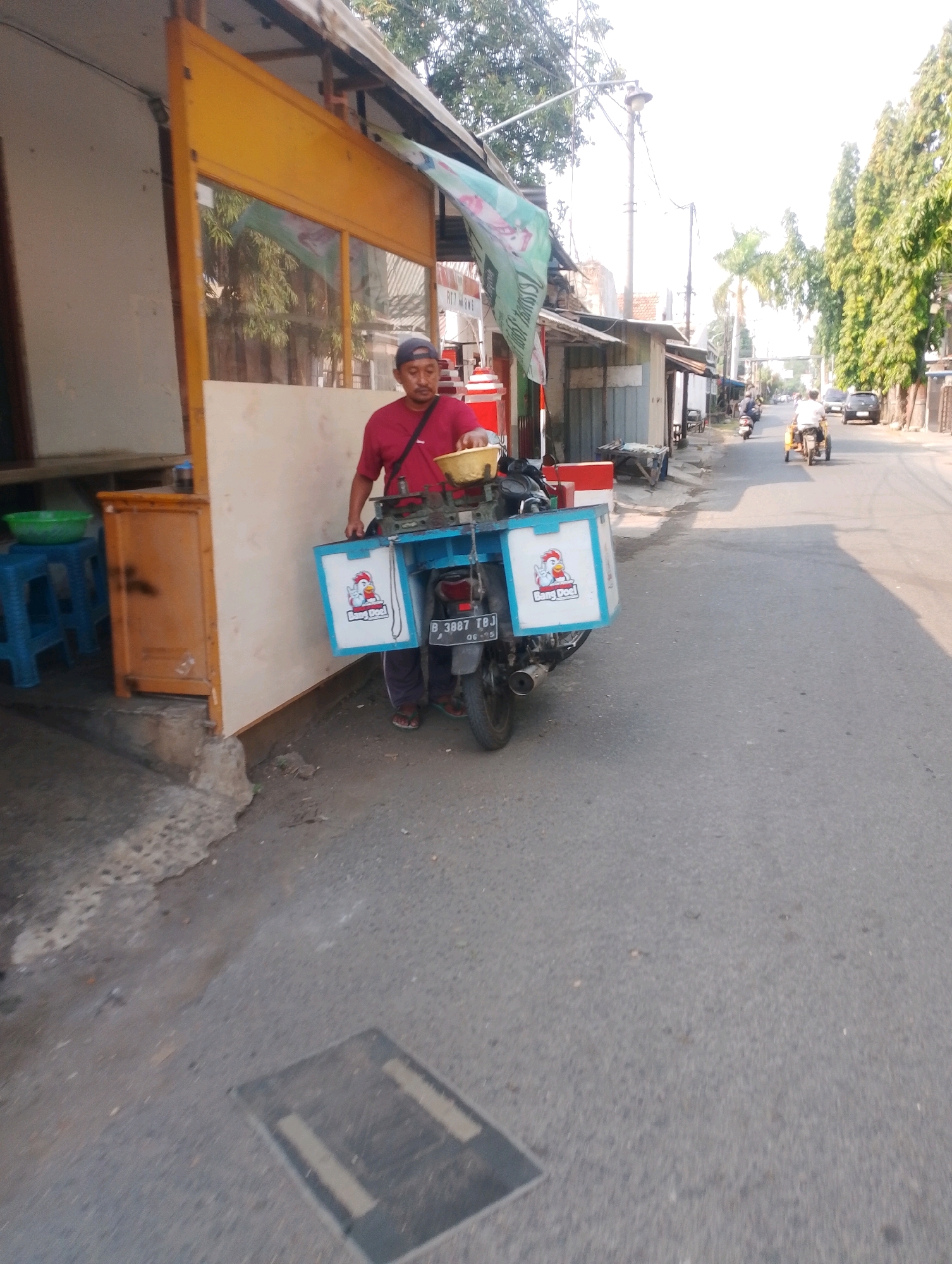 A man wearing a maroon t-shirt and a dark blue baseball cap stands next to a motorcycle in front of a food stall on a street in Probolinggo, Indonesia. The motorcycle has two white boxes attached to the rear, each with a cartoon chicken graphic and the text "Bang Doel" printed on them. The man is holding a yellow bowl in his right hand and appears to be preparing food. The food stall has a yellow awning and a wooden counter. In the background, other motorcycles and a car are visible on the street, along with trees and buildings. The scene depicts a typical street food vendor operation in a populated area of the city.