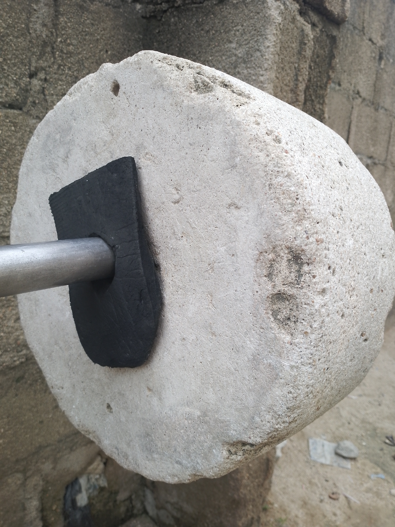 A close-up, eye-level shot shows a large, rough-textured, cylindrical grinding stone made of light-colored, porous rock. The stone is positioned horizontally, with its left side facing the camera. A dark, U-shaped piece of material, possibly rubber or thick felt, is attached to the front of the stone, surrounding a silver metal rod that pierces through the stone from left to right. The stone has a coarse, pitted surface with some darker inclusions and small holes. In the background, a rough, grey textured wall is visible, constructed of uneven blocks. The ground beneath the stone appears to be dirt and gravel. The lighting is even, with no strong shadows, suggesting an overcast day or indirect light.