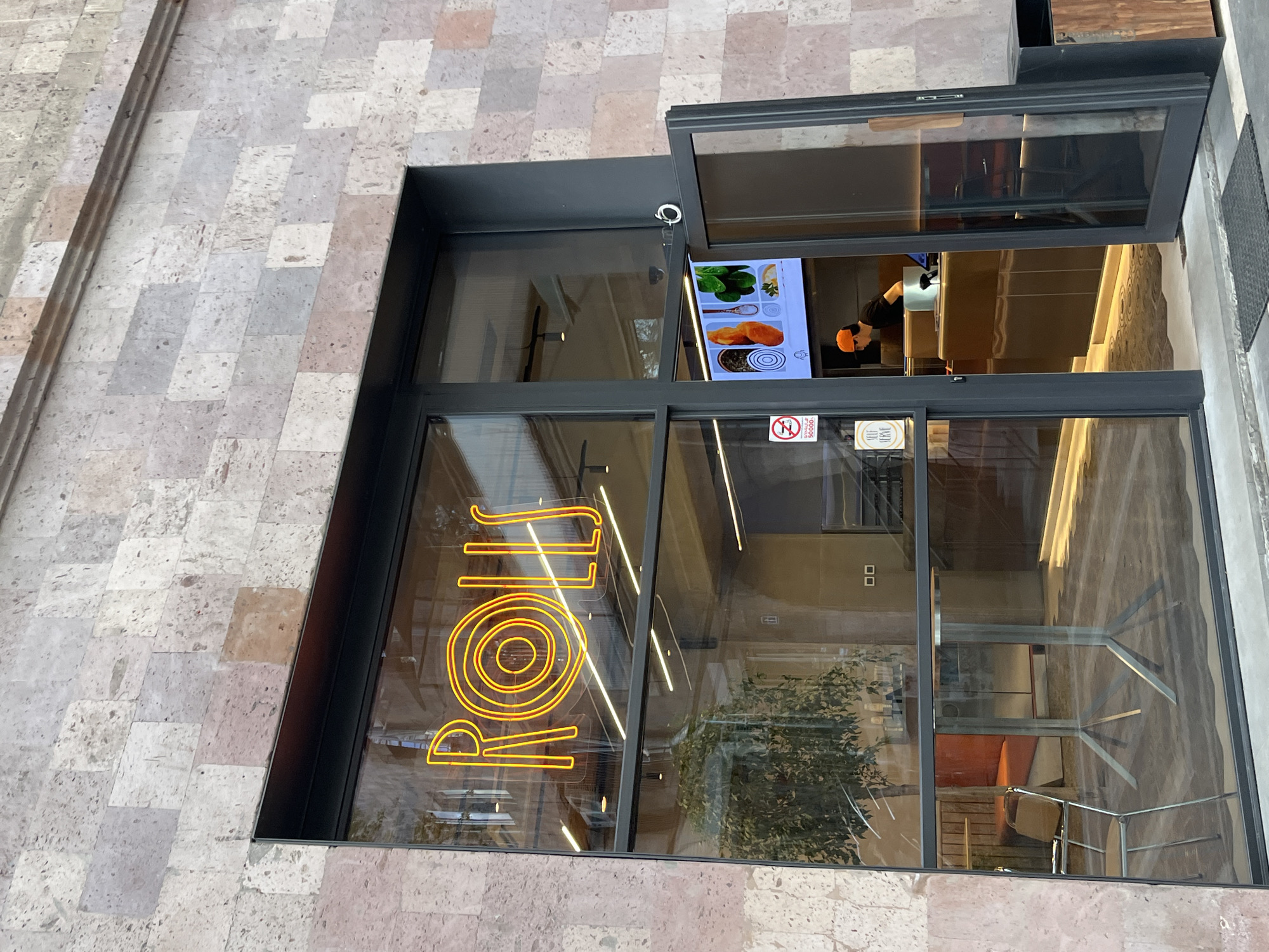 An eye-level, upward-tilted shot of a storefront with a light gray stone facade in Yerevan, Armenia. The sign on the storefront reads "ROLL" in yellow neon. A person wearing an orange hat can be seen inside. A menu screen is visible behind the sign. The interior of the store is visible, including tables and chairs. The storefront has a black frame, and the door above the entrance is open. A "No Smoking" sign is visible on the glass. The surrounding street and sidewalk are not directly visible but are hinted at by the angle of the shot.