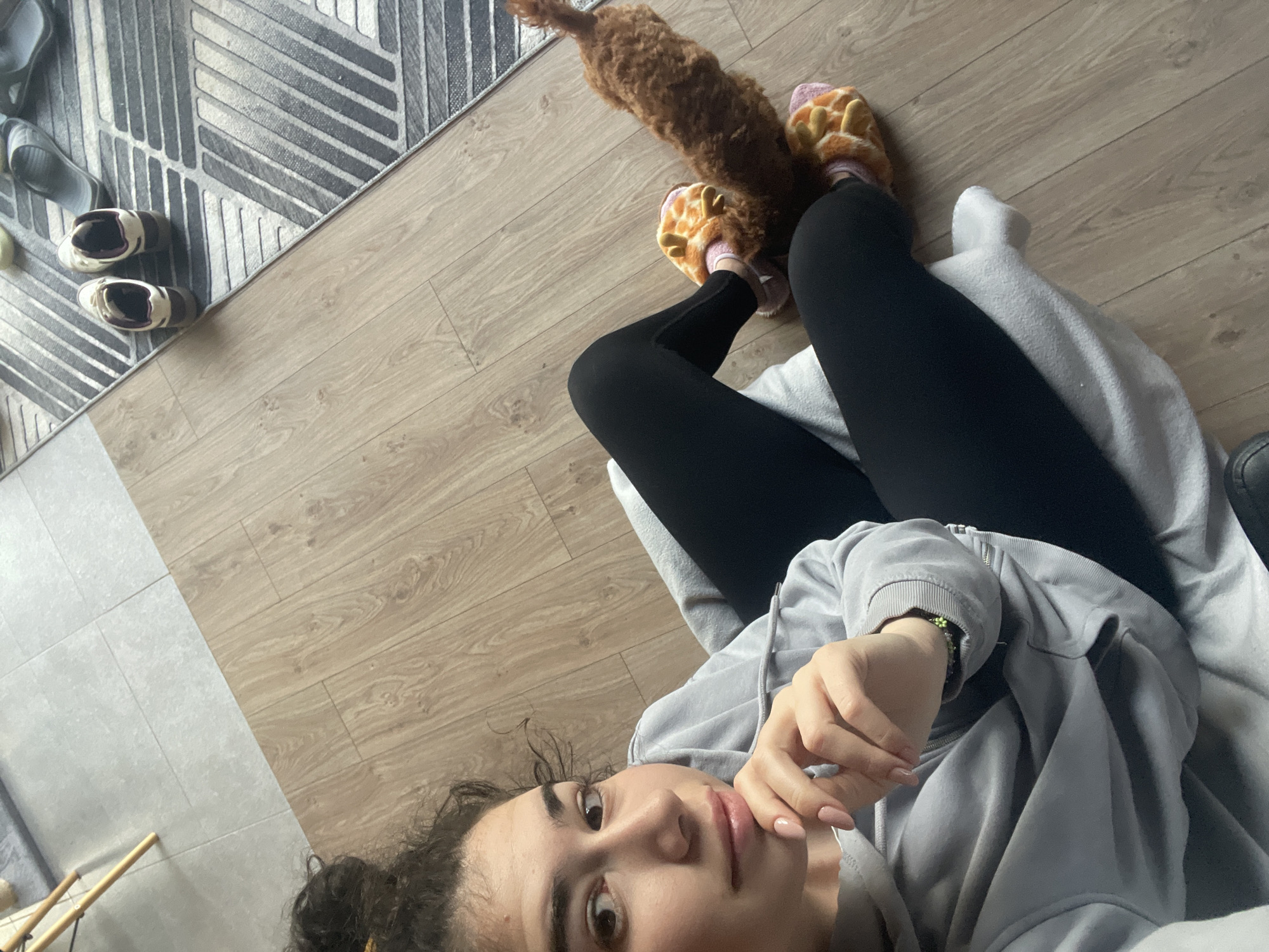 The image is a selfie taken indoors, likely from a high-angle perspective. A young woman with brown hair and fair skin is the primary subject, wearing a grey hoodie and black leggings. She is sitting on a light-grey blanket or cushion, legs slightly bent. On her feet, she wears orange giraffe-themed slippers. A brown, long-bodied dog is situated near her feet. The floor is light brown wood. In the background, a grey rug with a geometric pattern is visible, along with several pairs of grey/white shoes. Based on typical architecture and interior styles, the location appears to be a residential setting, but there is no evidence to pinpoint the exact location in Sofia, Bulgaria. The lighting suggests it's daytime. The woman is looking at the camera with a pensive expression, her hand near her mouth.