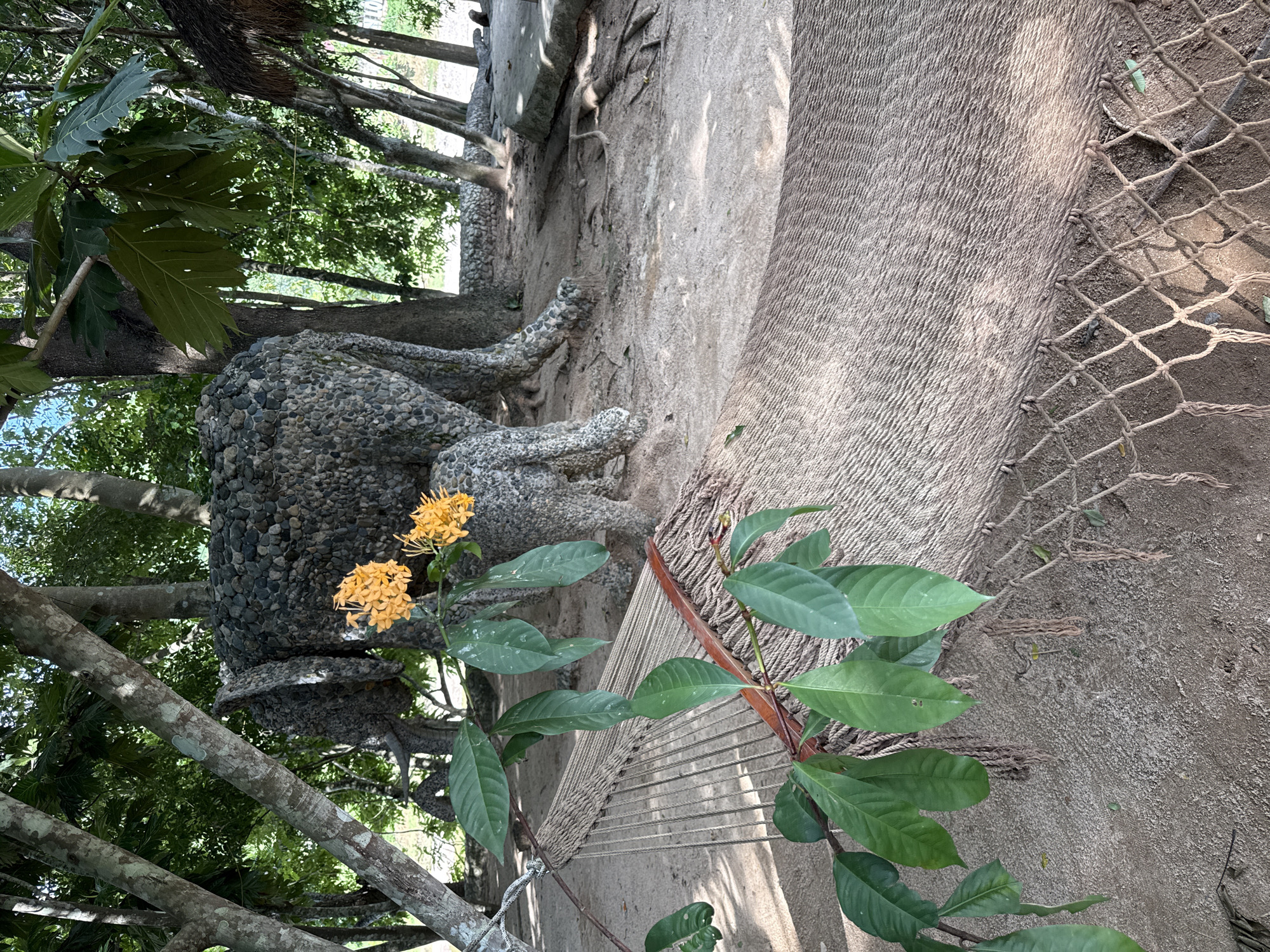 In a sandy, outdoor area with scattered trees and foliage, a hammock is strung between two trees. The hammock, made of tightly woven rope, is in the foreground, with a branch bearing green leaves and a cluster of small orange flowers extending over it. In the background, a large, textured sculpture resembling an elephant, constructed from dark gray and tan pebbles, stands amidst the trees. The elephant sculpture is partially obscured by tree trunks and branches. The ground is covered in fine sand and dirt, with visible tree roots. The lighting suggests it is daytime, with dappled sunlight filtering through the leaves. The location is identified as Nam Khanh Vinh city, Vietnam.