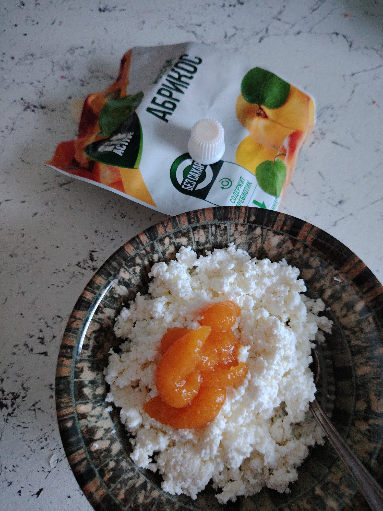 The image shows a close-up, overhead view of a bowl of cottage cheese topped with apricot slices, placed on a speckled white surface. To the upper right of the bowl is a pouch of apricot puree, with a white cap. The scene appears to be an indoor setting, possibly a kitchen counter. There are no people or animals present in the image. The lighting is natural, coming from above. The overall impression is one of a simple, healthy breakfast. There is no information in the image that would allow for the identification of the city of Vladimirovka, Russia.