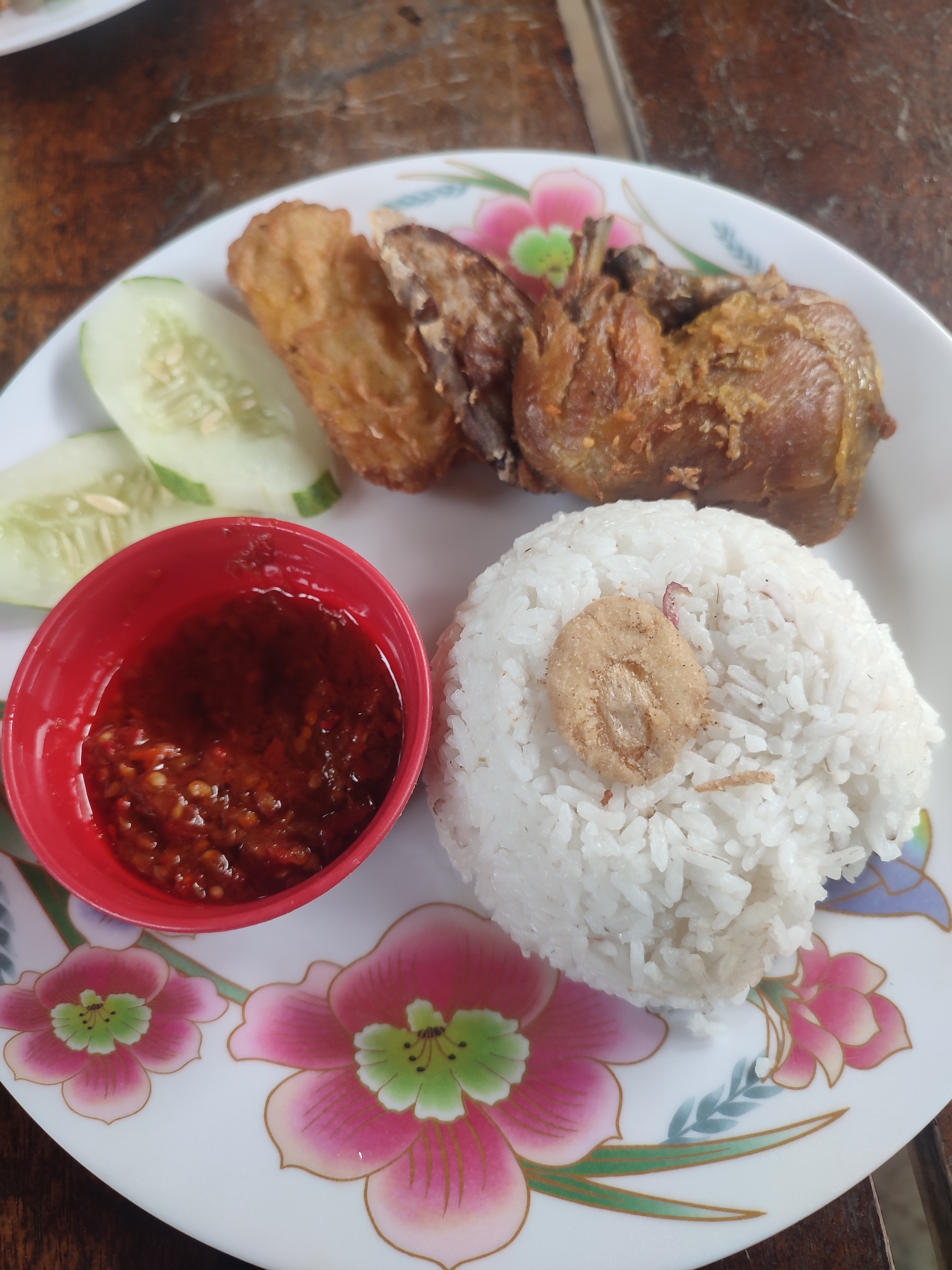 A close-up, high-angle shot captures a traditional Indonesian meal served on a white plate with a floral pattern, set against a blurred wooden table background. The main subjects of the meal are several pieces of golden-brown fried chicken, a mound of white rice, sliced cucumber, and a small red bowl of chili sauce (sambal).
The fried chicken, positioned towards the top right, exhibits a crispy texture and a deep, inviting brown hue. Two fresh, light green slices of cucumber are neatly arranged on the left side of the plate. Below the chicken and to the right is a generous serving of steamed white rice, topped with a single, oval-shaped piece of fried garnish, likely a fried shallot or garlic. The small red bowl, placed on the lower left, is filled with a thick, reddish-orange chili sauce, characteristic of Indonesian sambal, containing visible flecks of chili and spices.
The white ceramic plate is adorned with an intricate pattern of large, stylized pink and green flowers, adding a decorative element to the presentation. The wooden table surface in the background is out of focus, suggesting a rustic or casual dining setting. The meal's composition is typical of Indonesian cuisine, aligning with the location context of Keudealue Ie Puteh, Indonesia. There are no people visible, nor any discernible text. The time of day and weather cannot be determined from the image.