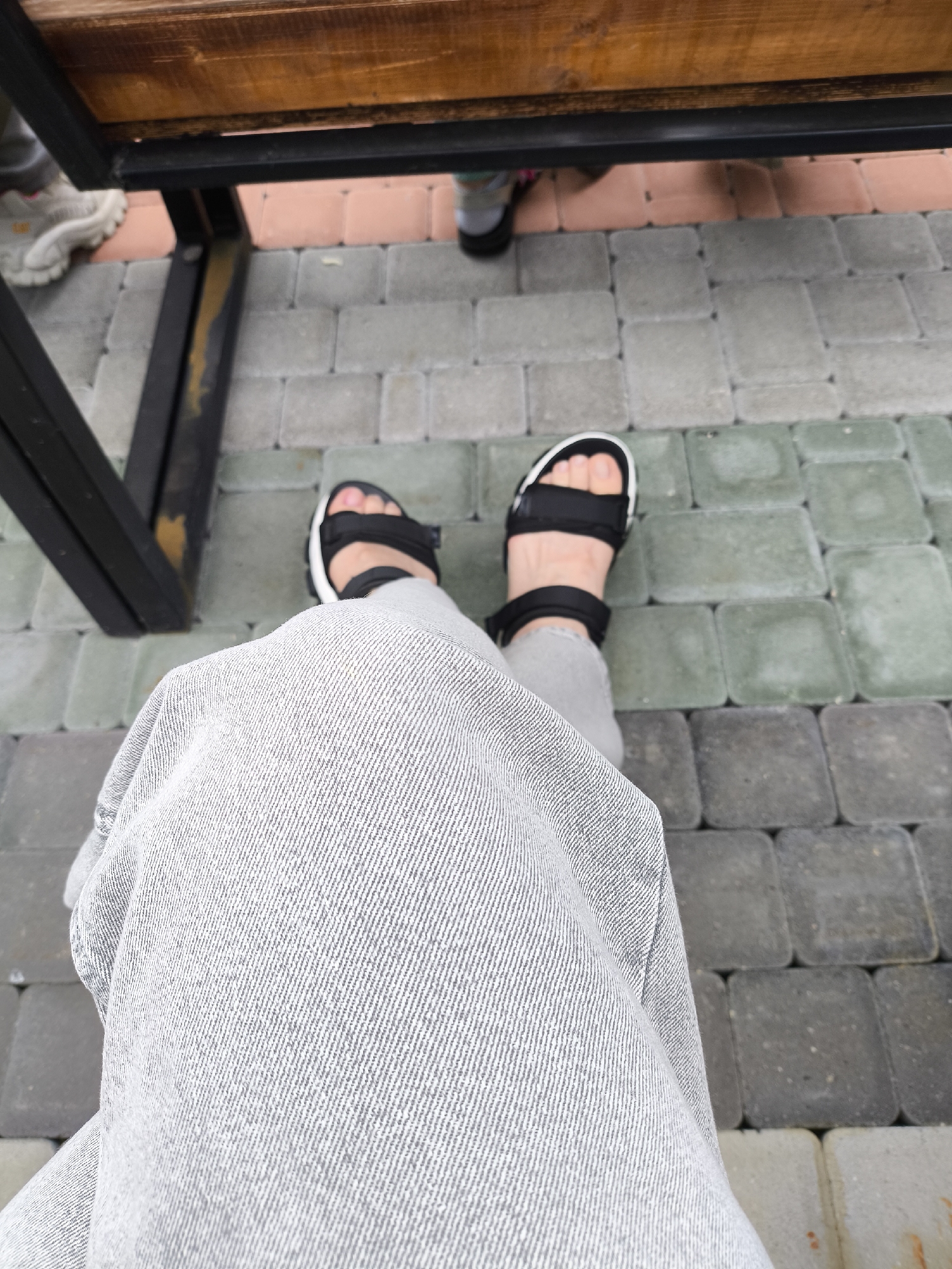 A first-person perspective shot shows a person's legs and feet clad in gray denim jeans and black-and-white platform sandals, resting on a paved ground composed of interlocking concrete pavers. In the background, the legs of other individuals are visible, some wearing athletic shoes, near a wooden bench supported by a black metal frame. The setting appears to be outdoors, possibly a park or an outdoor seating area, with the pavers indicating a walkway or patio. There are no overt indications that this image was taken in Borgustanskaya, Russia, or any other specific city.