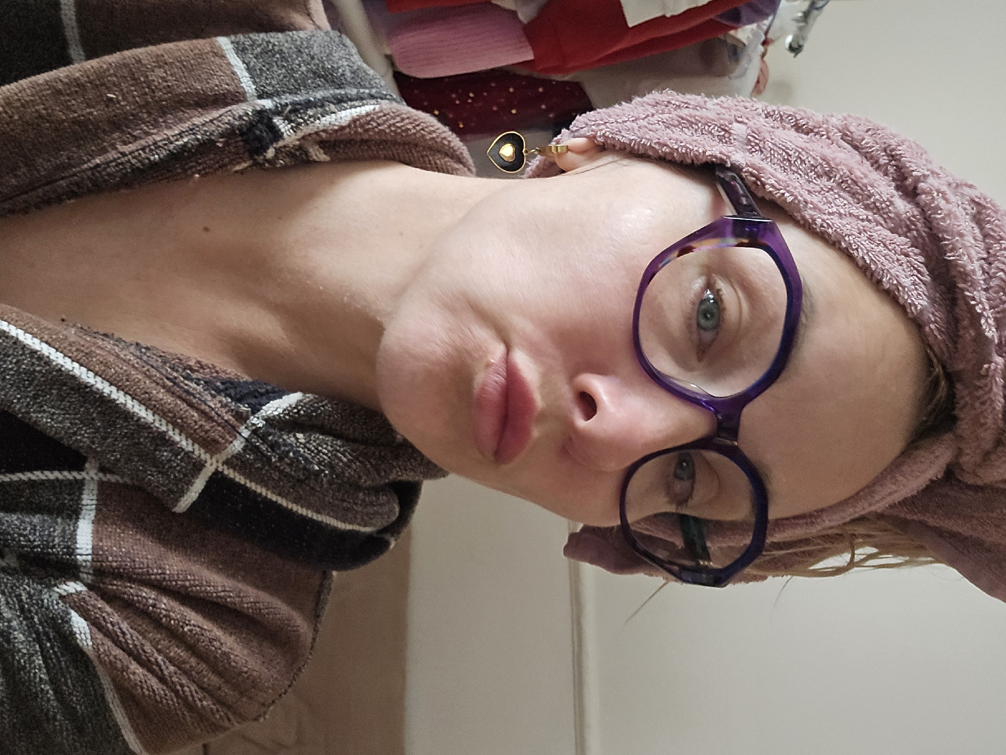 A person with fair skin, wearing purple eyeglasses, a pink towel wrapped around their head, and a brown and grey plaid robe, is looking at the camera. They are wearing a gold heart-shaped earring. The background includes a white wall, and some clothing stacked above and behind the person. The image appears to be taken indoors. There is no indication of Sochi, Russia.