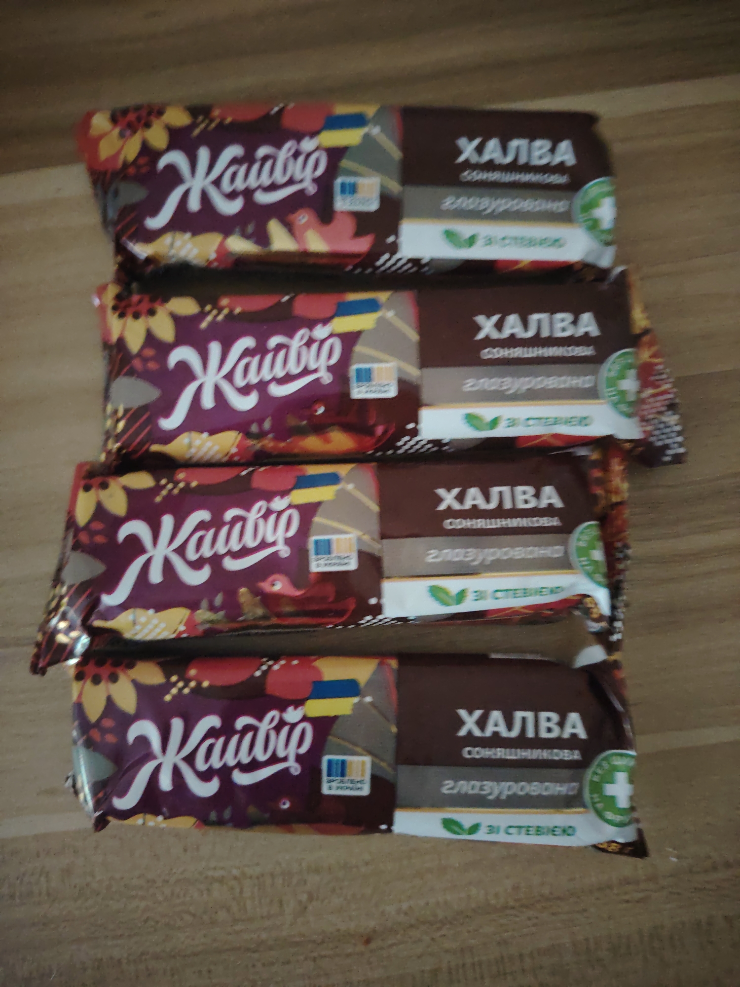 Four individually packaged halva bars are diagonally arranged on a light brown wooden surface, featuring visible grain. Each bar's packaging is bifurcated visually: the left section is dark purple with a brand name "Жайвір" in white script, adorned with illustrations of yellow sunflowers, red birds, and leaves. This section also includes a small Ukrainian flag graphic and a "ЗРОБЛЕНО В УКРАЇНІ" (Made in Ukraine) logo. The right section is dark brown, displaying "ХАЛВА" (Halva) in prominent white uppercase letters, followed by "СОНЯШНИКОВА" (Sunflower), "ГЛАЗУРОВАНА" (Glazed/Coated), and "ЗI СТЕВIЕЮ" (With Stevia) next to a green leaf icon. A green circular emblem with a white plus sign is present on the far right of this section. The bars are slightly staggered, appearing to overlap, with the bottom bar positioned lower and to the left of the others. No individuals are present in the scene, and no actions or interactions are depicted.