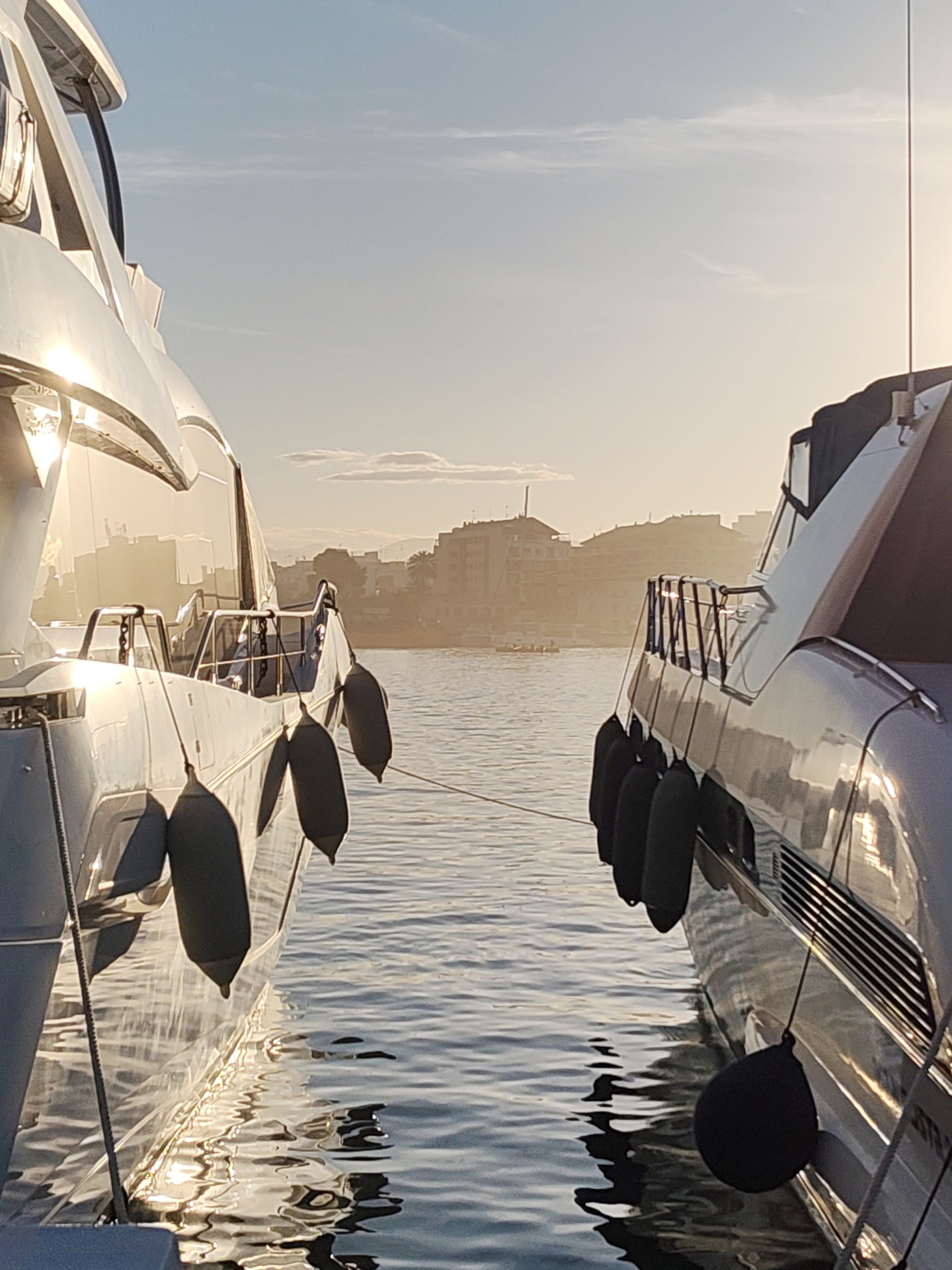 The image depicts two luxury yachts moored in Dénia, Spain, likely within the port or marina, as suggested by the urban backdrop visible across the water. The yachts are positioned closely, with fenders hanging between them to prevent damage. A mooring line connects the two boats. The city buildings, slightly obscured by haze or mist, rise in the background. The time of day is likely morning or late afternoon due to the low angle of the sunlight reflecting off the hulls of the yachts. The water is calm, reflecting light and shadow, and the overall atmosphere is serene. There is a small boat in the distance.