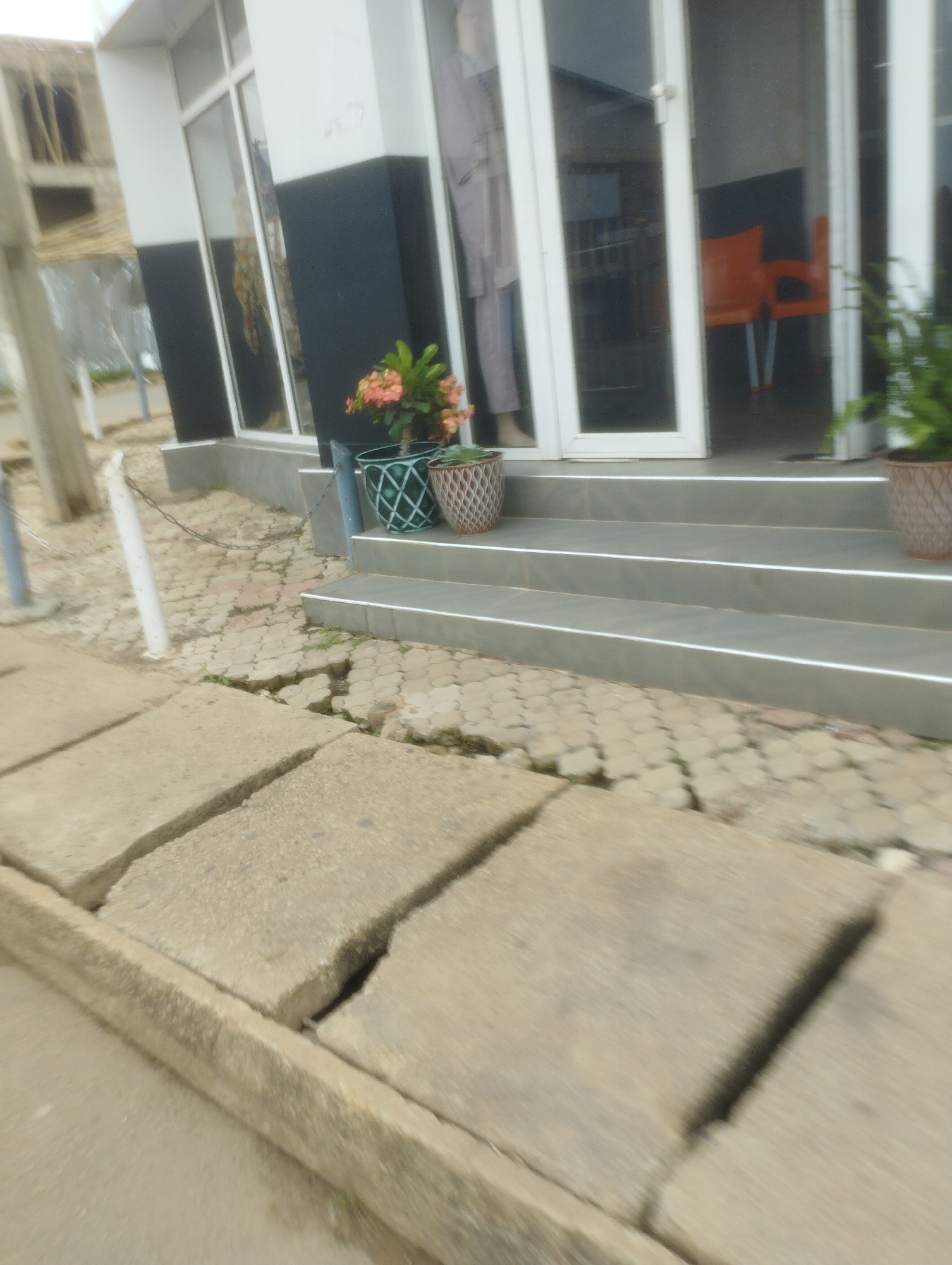 The image depicts a street scene in Jos, Nigeria, with a focus on a building's entrance. In the foreground, cracked concrete slabs form a pathway, showing signs of disrepair. A low concrete curb borders this path. Further back, a set of grey tiled steps leads up to the entrance of a building. The building features a large glass-fronted door with white frames. Through the glass, two bright orange chairs are visible inside.
To the left of the entrance, a plant with small pink flowers in a decorative pot sits on the ground. To the right, a large green plant in a similarly patterned pot is visible. The exterior wall of the building is split horizontally, with the upper section painted white and the lower section a dark color, possibly black. Behind the building, some construction or scaffolding is visible. A metal chain is strung between two posts on the left side of the image, near the building's entrance. The ground around the building's base is paved with interlocking stones, which also appear to be uneven and somewhat damaged in places. The overall impression is of an urban environment with some areas experiencing structural wear and tear.