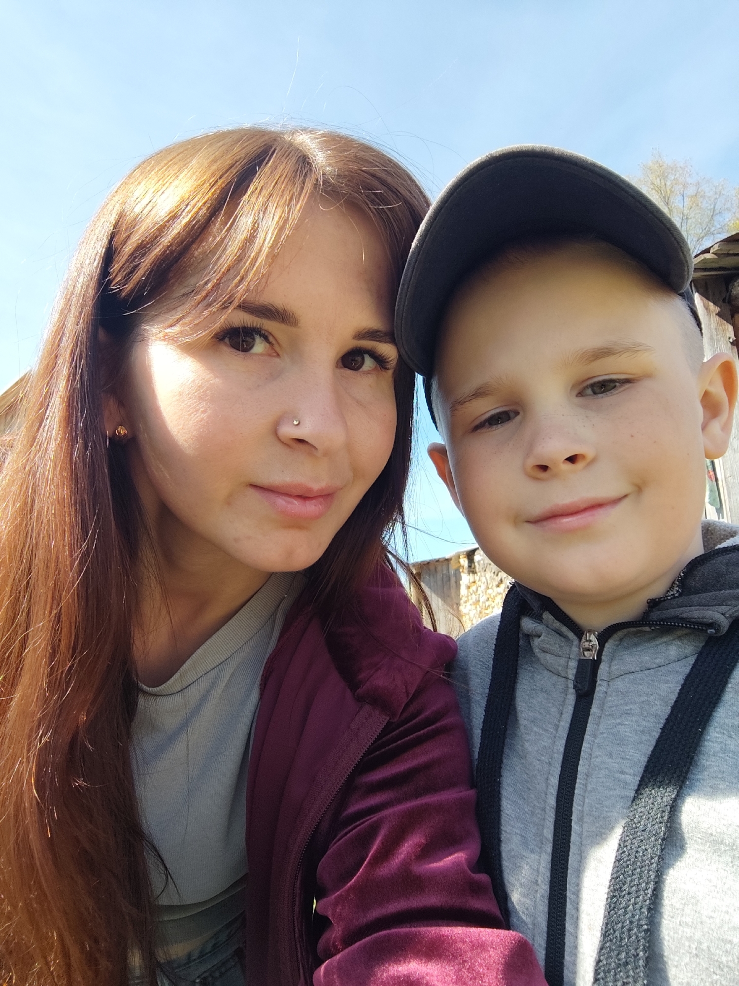 The image depicts a selfie of two individuals: an adult female and a young boy. The female has long, reddish-brown hair with bangs, fair skin, and a nose piercing. She is wearing a grey top and a maroon velvet jacket. The boy has fair skin, short blond hair, and is wearing a baseball cap and a grey hooded sweatshirt. The background appears to be outdoors, with a blue sky visible above. Weathered wooden structures are visible behind the subjects. The location might be in Фоки, Russia, given the prompt's instructions.