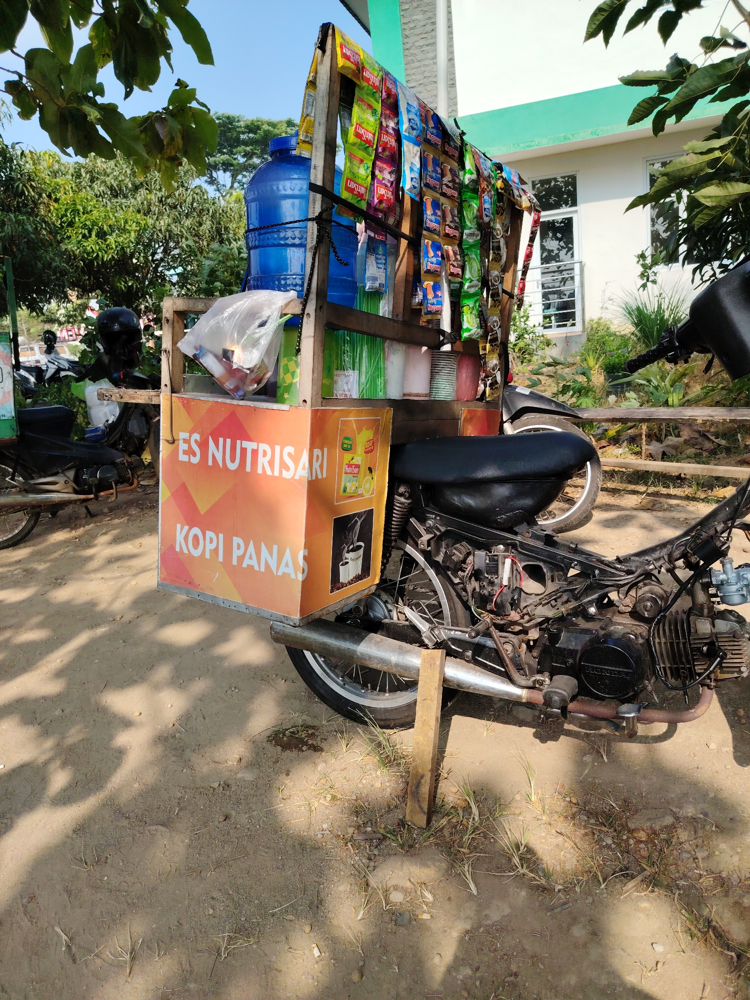 This is a medium shot of a mobile beverage cart attached to a motorcycle, parked on a dirt path in front of a building. The cart is constructed of wood and metal, with a prominent orange panel displaying "ES NUTRISARI KOPI PANAS" in white lettering. Various sachets of beverages, including Nutrisari and possibly coffee, are hung on the side of the cart. A large blue water jug is visible at the top of the cart, along with a plastic bag containing items. The motorcycle itself is partially disassembled, with visible engine components and wiring. The scene is outdoors, with sunlight casting shadows on the ground. Lush green trees and foliage are visible in the background, along with a white building with green trim and windows. The overall setting appears to be a roadside or informal marketplace in a tropical climate, consistent with urban or suburban areas of Semarang, Indonesia. There are no people visible in the frame.