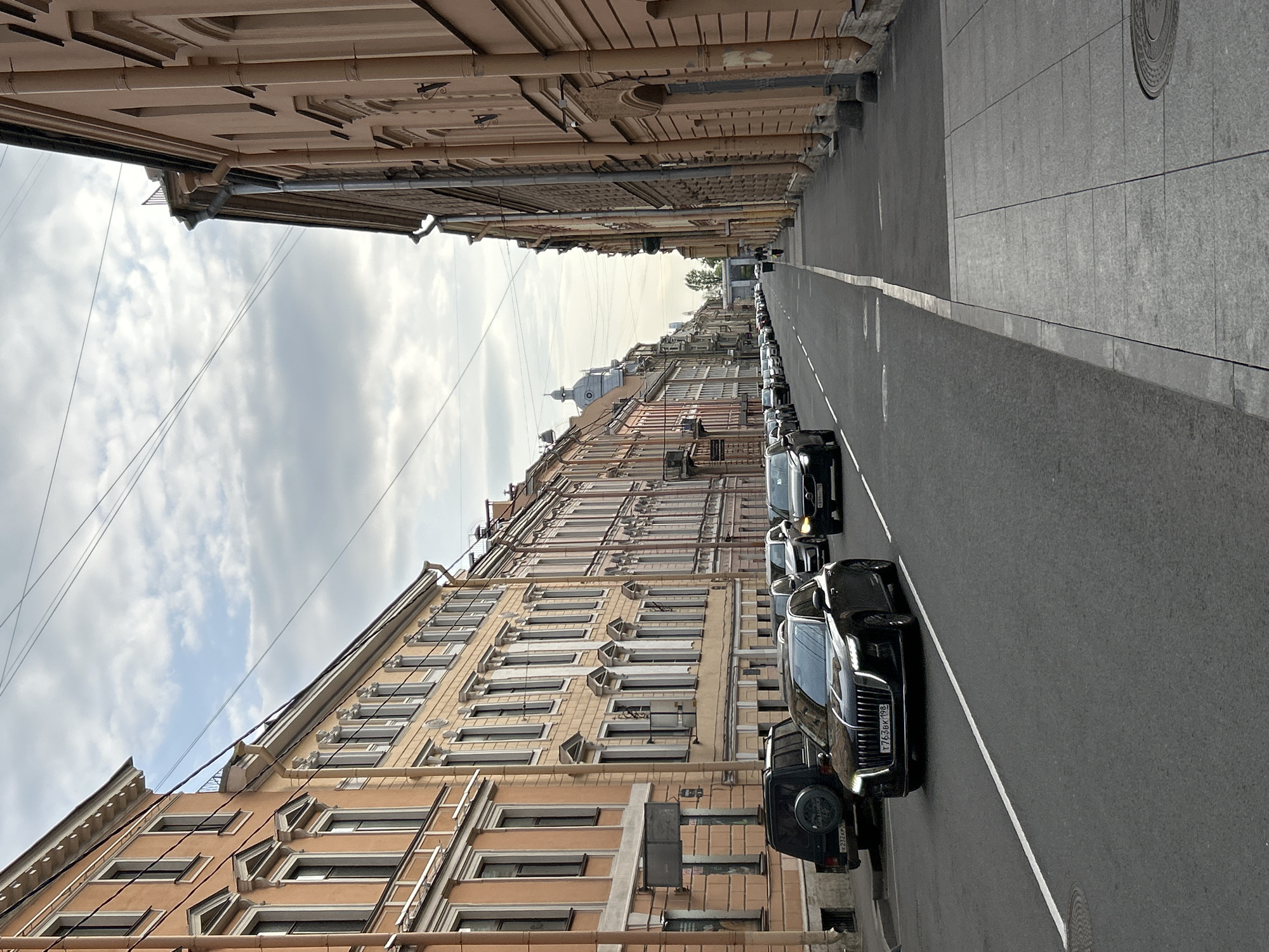 The image captures a street in Saint Petersburg, Russia. A line of dark-colored vehicles is parked along the right side of the road, adjacent to the sidewalk. The road is paved, marked with a white line dividing traffic lanes. Tall buildings line both sides of the street, showcasing classic architectural details with multiple windows and intricate facades. The sky is overcast, with grey clouds. Power lines are visible stretching across the sky between buildings. The overall scene depicts an urban environment, likely a central area within Saint Petersburg.