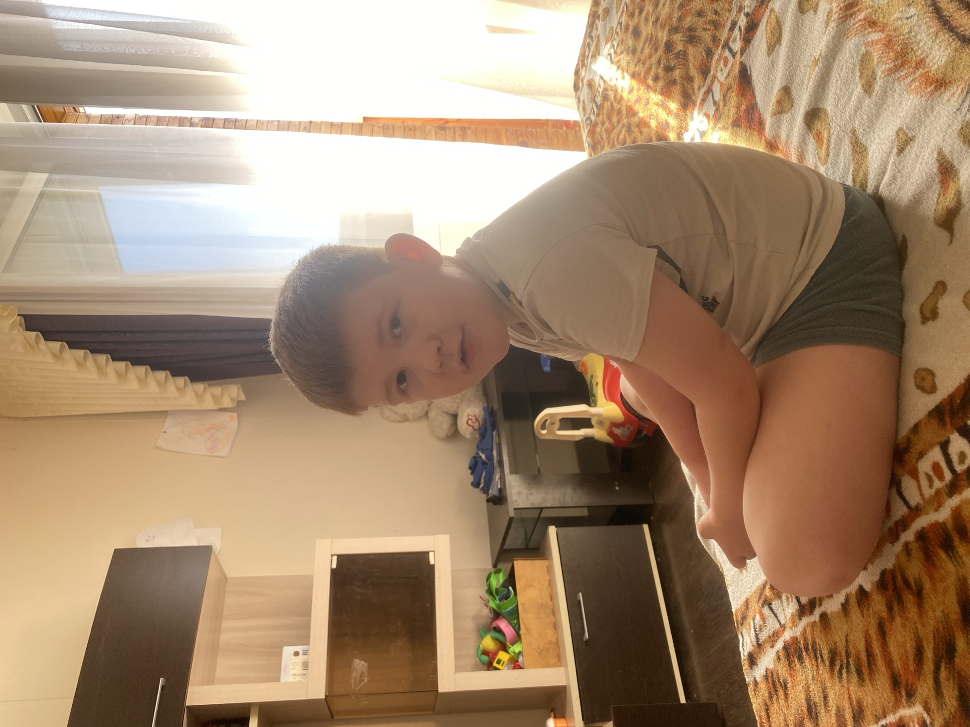 A child sits on a bed in a room. The child is fair-skinned, with short brown hair, wearing a beige t-shirt and gray shorts. The bed has a patterned brown and white cover. Behind the child is a shelf with a closed cabinet, a toy motorcycle, and a collection of colorful toys. Above the shelf, a child's drawing is taped to the wall. A window covered with light-colored curtains is visible in the background. Based on the image's metadata, the photo was taken in Chaikovsky, Russia.