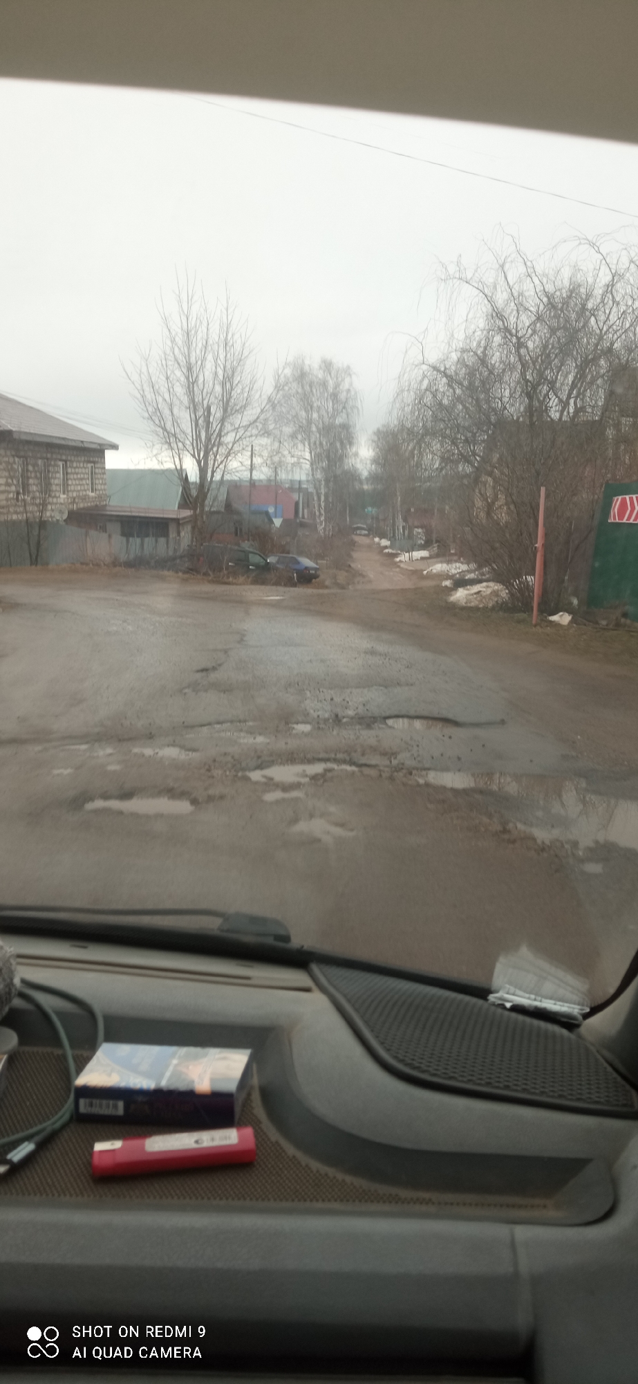 The image shows a view from inside a vehicle, likely a car or truck, looking out onto a street in a residential area. The location appears to be in a city called Unknown, within an unknown region. The street is heavily potholed and contains puddles, suggesting recent rain or snow melt. Houses are visible on both sides of the street, with bare trees and remnants of snow indicating it may be late winter or early spring. There are two dark cars parked on the left side of the road. The dashboard of the vehicle is visible in the foreground. A pack of cigarettes and a red lighter are on the dashboard. The sky is overcast, contributing to the muted colors of the scene.