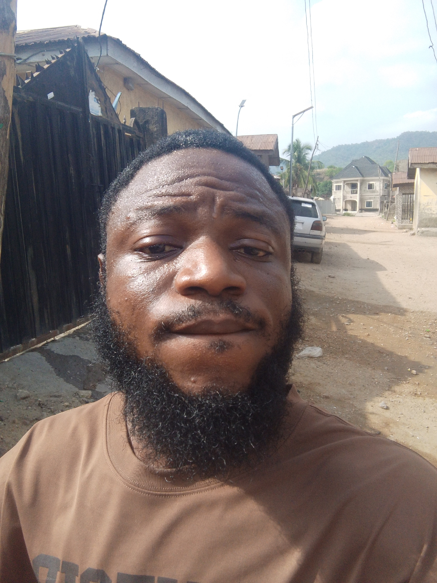 A selfie-style, close-up portrait features a man with dark skin, a full black beard, and short, dark, kinky hair, looking directly into the camera. A noticeable sheen on his forehead and face suggests warmth or humidity. He is wearing a plain brown short-sleeved t-shirt, with some dark, illegible text visible on the lower right side.
The background portrays an outdoor, semi-urban environment in Lokoja, Nigeria, bathed in bright daylight. To the man's left, a dark corrugated metal fence or gate stands beside a building featuring a similar roof. To his right, a dusty, unpaved road extends into the distance, with a light-colored SUV parked along its side. Further down the road, a two-story building with a dark roof and other structures are visible. Overhead, power lines traverse a clear, bright sky, along with two street lights. In the far distance, lush green hills or mountains form the horizon, accompanied by scattered palm trees and other vegetation. The scene evokes a sunny and likely warm day in a residential area.