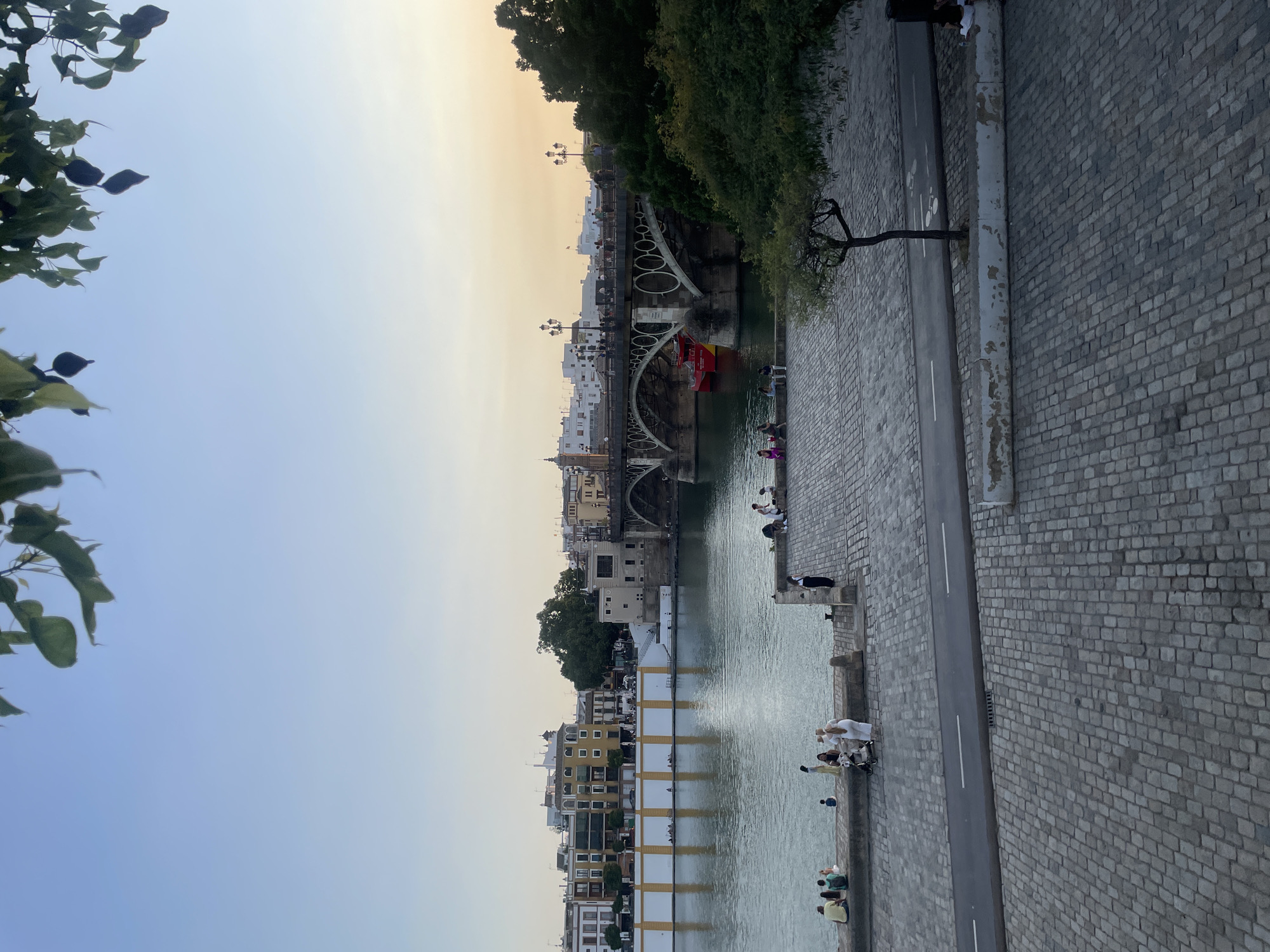 The image depicts a view of the Guadalquivir River in Seville, Spain, with the Triana Bridge in the background. The scene is set during the late afternoon or early evening, as indicated by the soft, warm light and the clear sky.
In the foreground, a cobblestone walkway lines the riverbank, with several people seated or standing, enjoying the view. Some are casually dressed, with a few couples and small groups visible. A baby stroller is present, suggesting families are present.
The river itself is calm, reflecting the buildings and the sky. Along the opposite bank, there are several buildings with a distinctive yellow facade, some with white detailing. Further down the river, the Triana Bridge, an iconic iron bridge with multiple arches, spans the water. People can be seen walking across the bridge, and a bright red boat is docked beneath one of its arches.
To the right of the image, a lush green tree with a distinctively shaped trunk stands near the walkway. The overall atmosphere is peaceful and relaxed, capturing a common scene of leisure and city life in Seville. The image is taken from a slightly elevated perspective, looking down onto the river and the bridge.