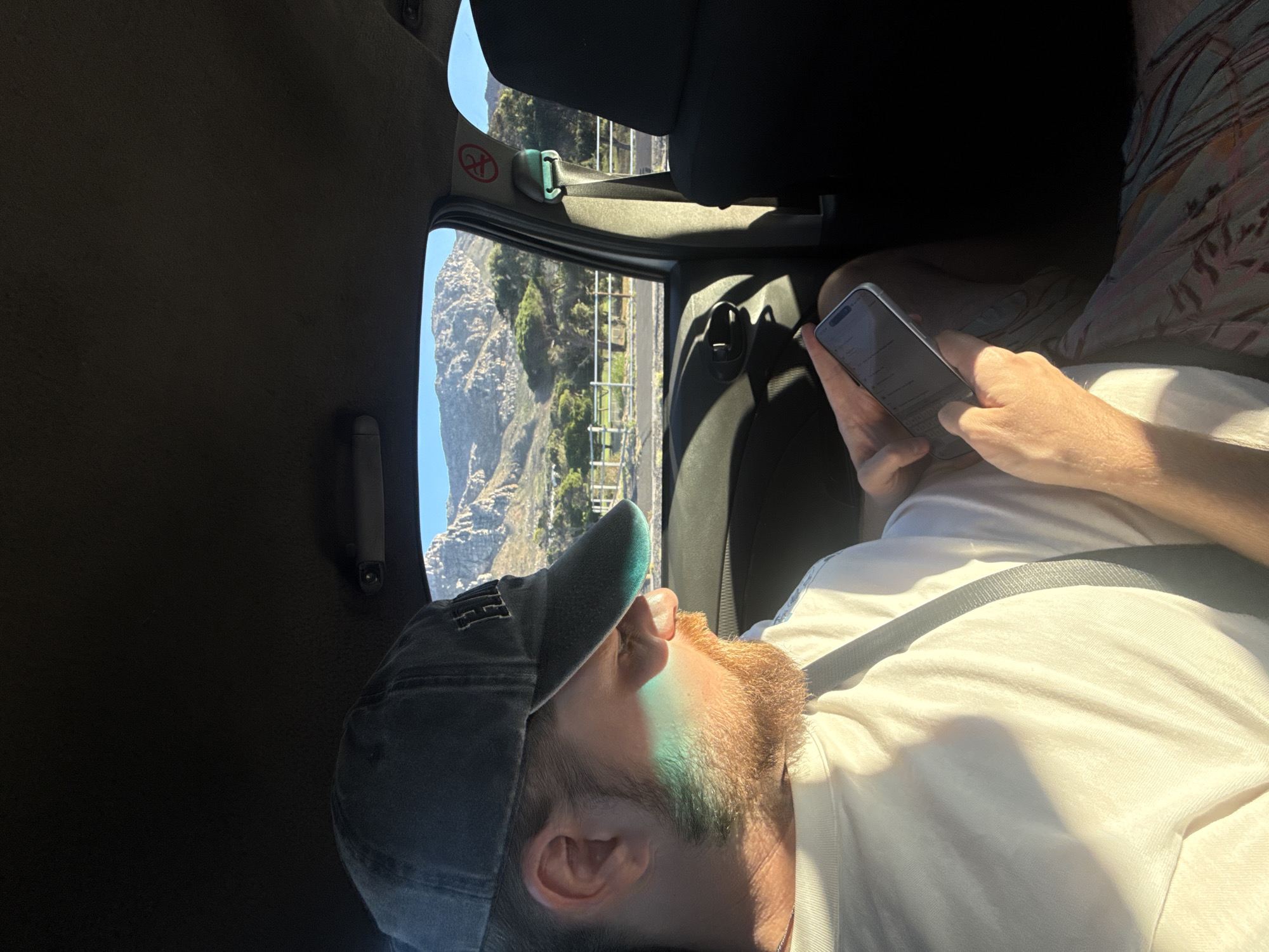 A man wearing a cap and a white t-shirt is seated in a vehicle, looking at his phone. He is wearing a seatbelt. The window shows a view of a mountain alongside a road, indicating that the vehicle is likely moving. Based on the landscape and the light conditions, the location is likely in the vicinity of Cape Town, South Africa, possibly on a scenic route along the coast near a mountain range. The vehicle's interior is dark.