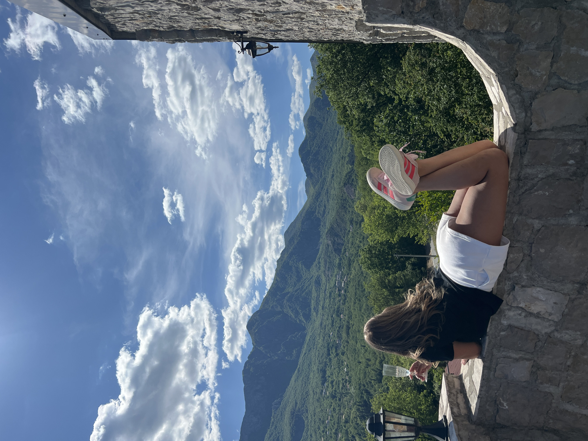 A rotated outdoor shot shows a young woman sitting on a stone structure. She is seen from the back, wearing a black shirt and white shorts. Her bare legs are raised towards the green forested mountain and blue cloudy sky in the background. She is wearing pink Adidas sneakers with white soles. She appears to be holding a crystal glass, partially obscured. The stone structure is likely a wall or archway, suggesting a scenic overlook. A vintage-style lamp is positioned on the stone structure near the woman. Sotonići, Montenegro cannot be ascertained from this photograph.