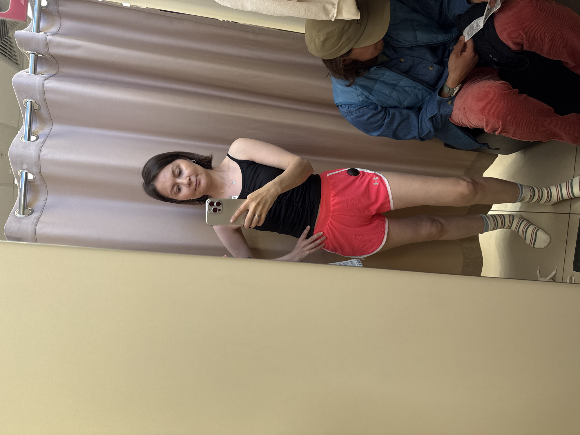 A woman is taking a selfie in a dressing room. She is wearing a black tank top and red shorts. She is holding a phone in her right hand and looking at the screen. There is another person seated in the dressing room behind her. The dressing room has beige walls and a curtain with silver hooks. It appears to be located in Izhevsk, Russia.