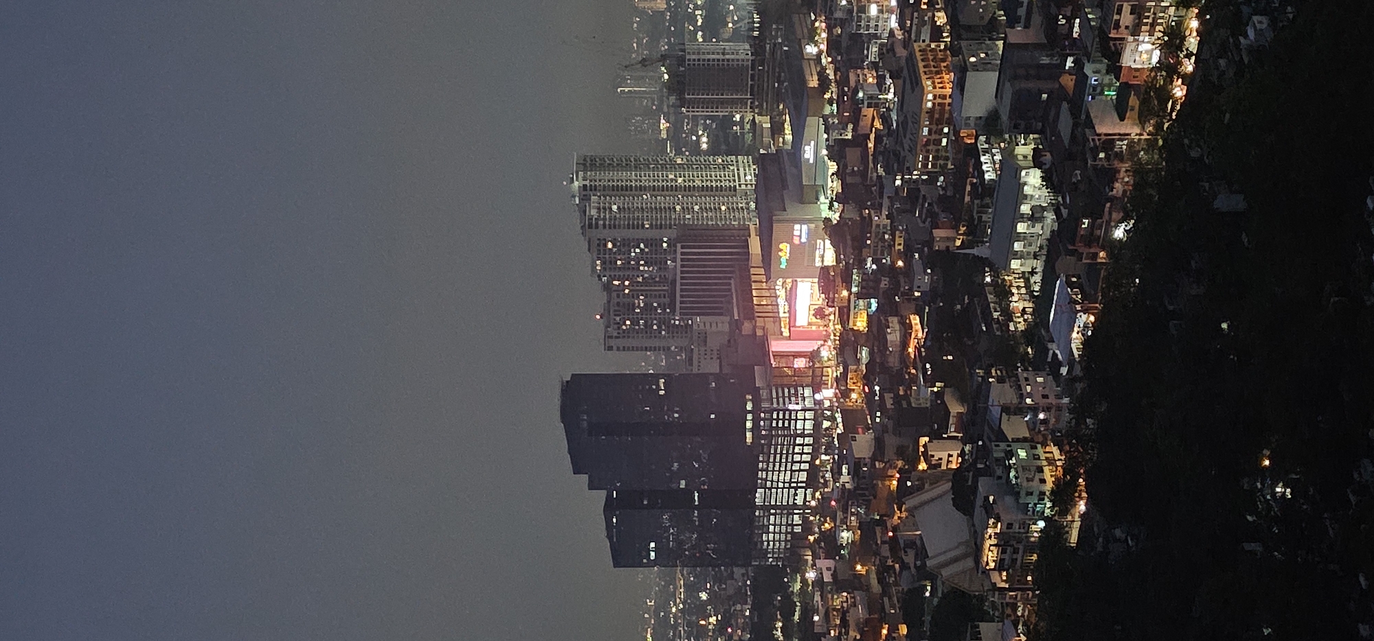 This is an aerial, nighttime photograph of the cityscape of Makati, Philippines. The scene captures a dense urban environment with numerous buildings illuminated by artificial lights. In the mid-ground, a large commercial complex with bright signage stands out, possibly a shopping mall or entertainment center. This complex is surrounded by various buildings of different heights and architectural styles, including prominent high-rise buildings and lower-rise residential or commercial structures. The overall impression is a bustling city at night, with the distinct glow of lights indicating economic activity and residential presence. The foreground is dominated by a dark, vegetated area, possibly a hillside or park, with scattered lights suggesting a cemetery or park amenities. The sky is dark and hazy, typical of a polluted urban atmosphere at night, with a faint gradient of lighter tones towards the horizon.