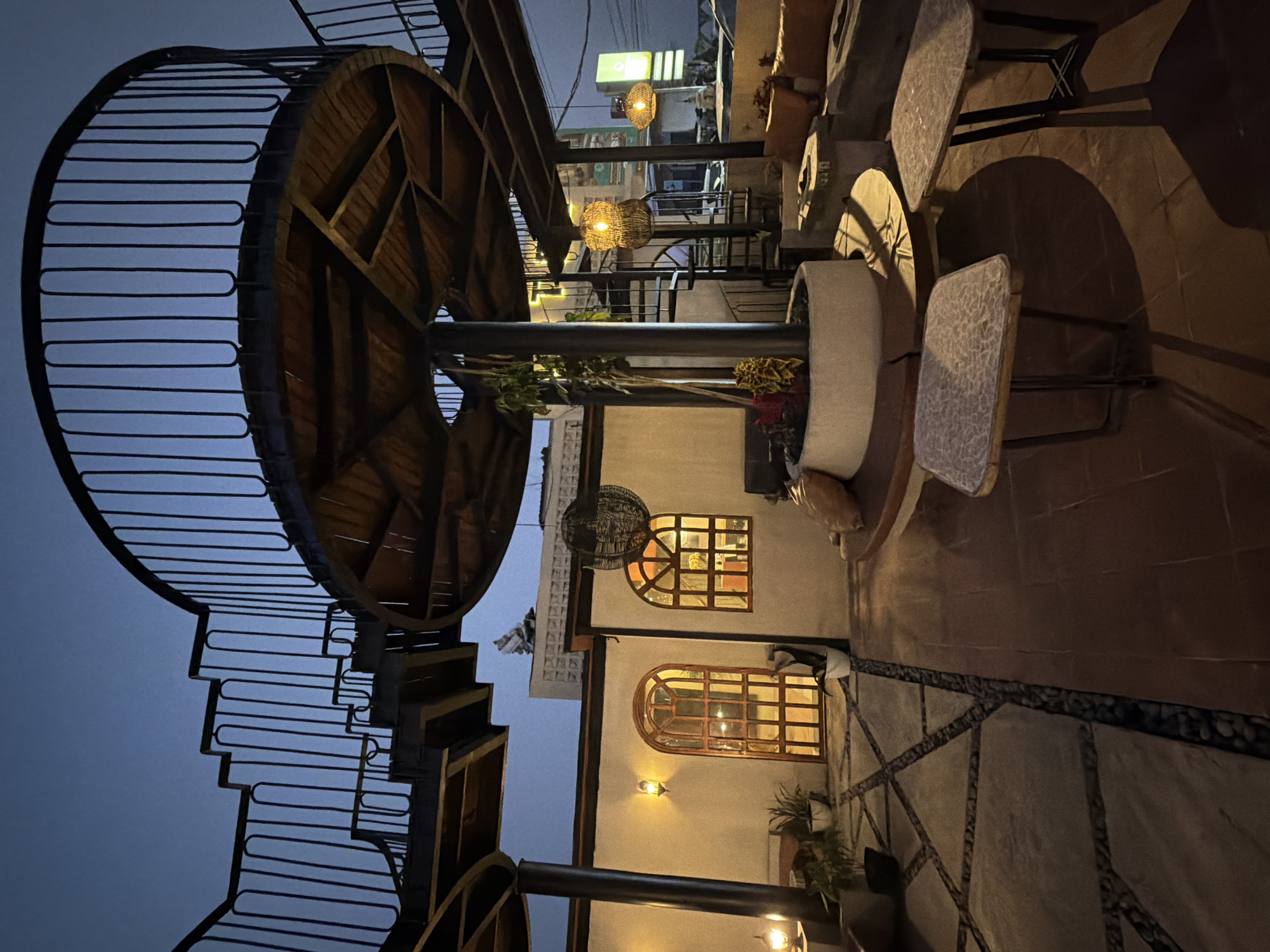The image shows an outdoor seating area at what appears to be a restaurant or cafe at dusk. A spiral staircase made of metal and wood is prominently featured on the left, leading up to a circular wooden structure. The seating area includes several tables and chairs, some with patterned cushions. One large, cylindrical object, possibly a planter or decorative element, is positioned near the tables.
The setting is a rooftop or elevated area, with views of buildings and lights in the background. Two woven pendant lights hang from above, casting a warm glow. The architecture features arched windows with wooden frames, and the walls are painted a light color. Potted plants are visible, adding greenery to the scene. The ground is paved with tiles and decorative stones.
The time of day is evening, as indicated by the dim natural light and the artificial lighting from the lamps and buildings. There are no people visible in the image, suggesting the area is either closed or the patrons are not in the frame. The overall mood is quiet and inviting, with the warm lighting creating a cozy atmosphere. No text is visible in the image. The location context of Kintamani, Indonesia, suggests a potentially scenic area, though the background details are not clear enough to confirm this.