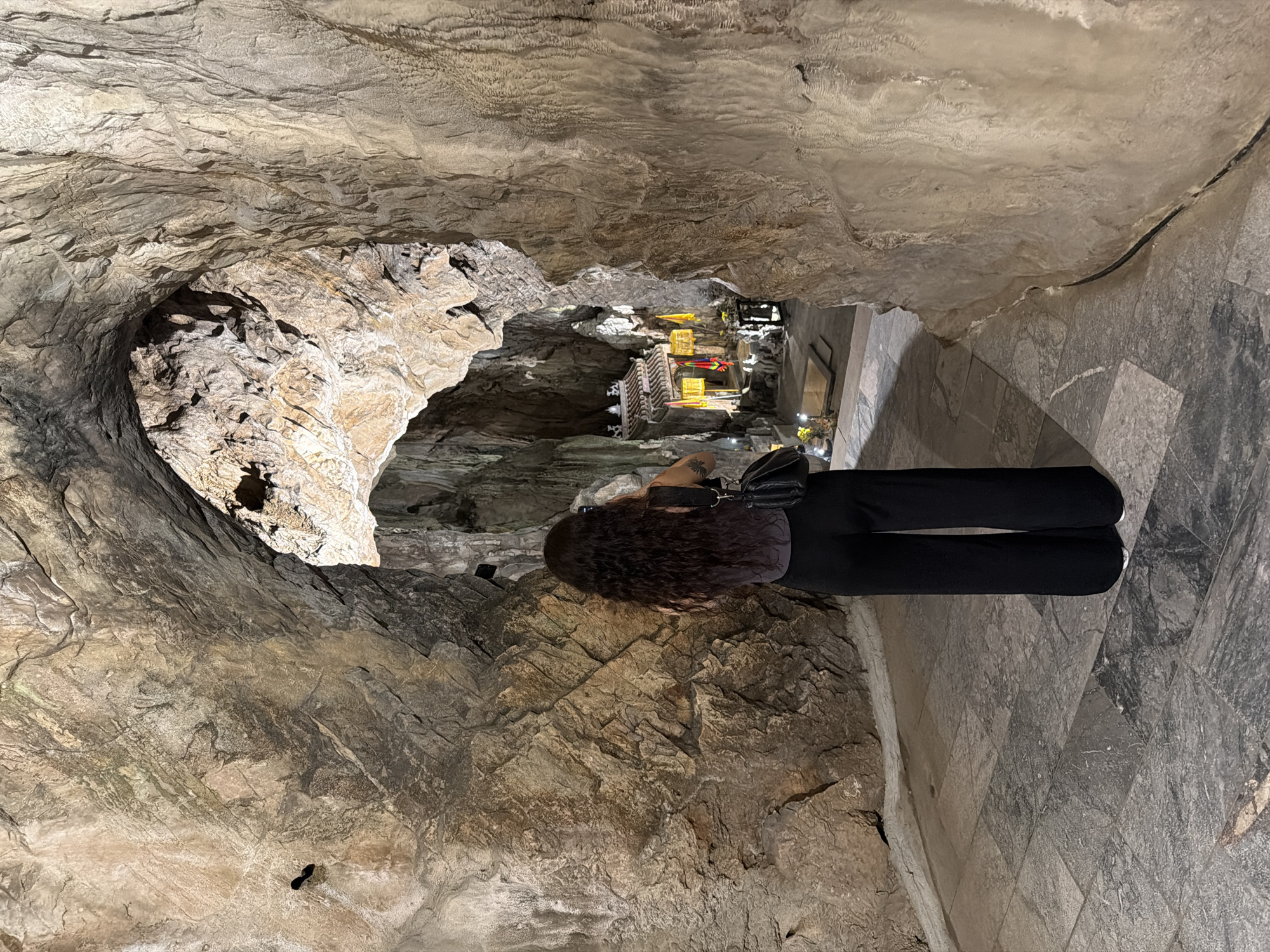 A woman stands at the edge of a cavernous opening, her back to the viewer, facing into a cave adorned with lighting and possibly structures. She wears dark pants, a dark top, and has dark, curly hair. A black bag hangs from her shoulder. The cave is rocky and textured with shades of beige, brown, and grey. A passageway with tile flooring is adjacent to the cave opening. Based on the location, the image appears to be taken in the Marble Mountains caves in Đà Nẵng, Vietnam.