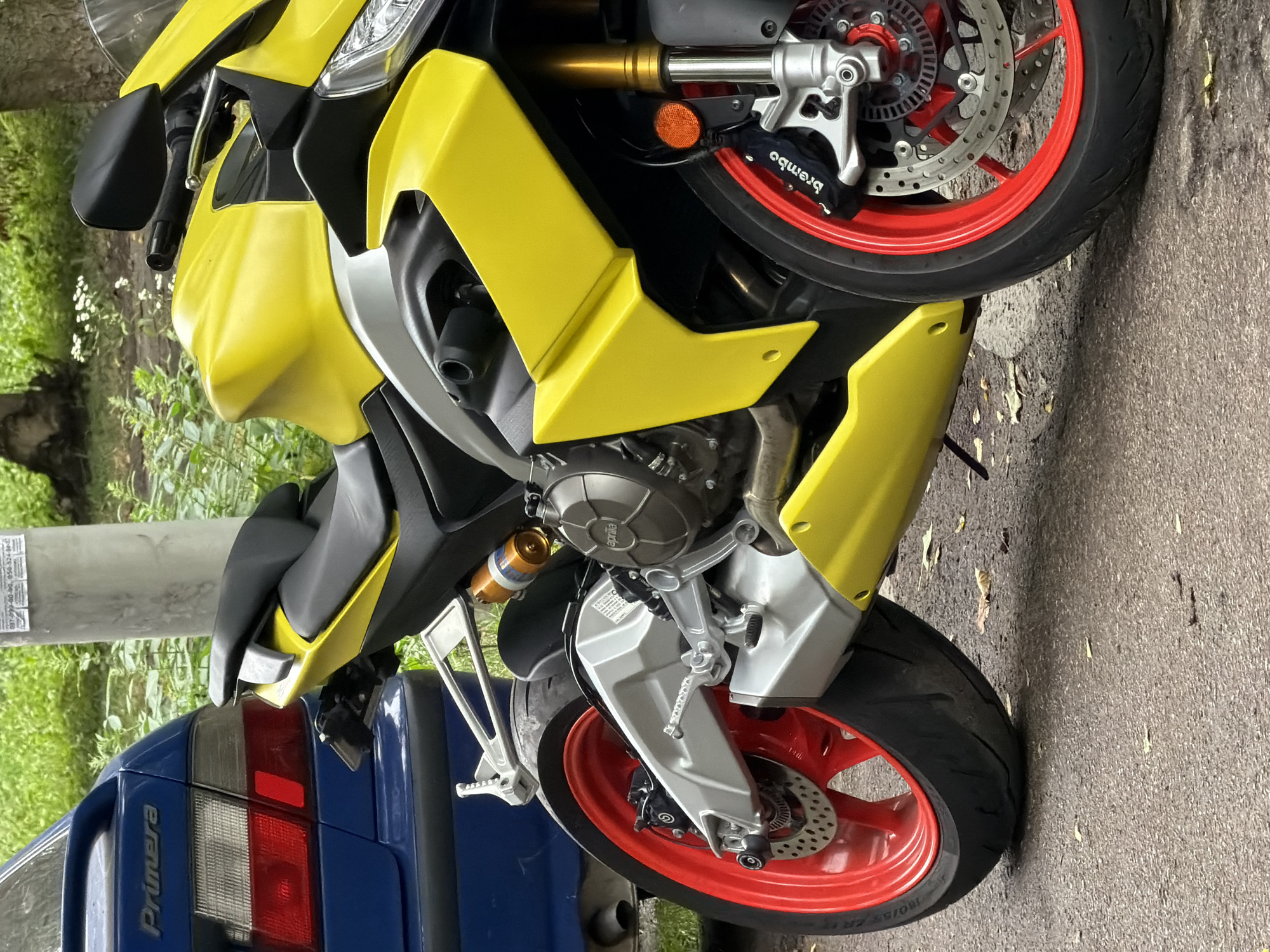 A yellow and silver sport motorcycle is parked next to a blue car on a paved surface, likely a street or parking lot. The motorcycle's rear wheel has a red rim and is fitted with a disc brake and caliper labeled "Brembo." A portion of the motorcycle's engine, exhaust, and shock absorber are visible. The blue car appears to be a Nissan Primera, indicated by the "Primera" emblem on its trunk. The background consists of greenery, including trees and foliage, and a concrete utility pole. The scene is outdoors, and the lighting suggests it is daytime. The image was taken in Kyiv, Ukraine.