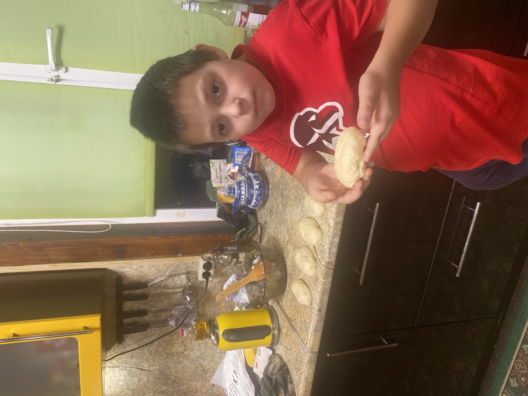 A young boy is standing in a kitchen, holding a piece of dough. He is wearing a red t-shirt with a white graphic. In front of him on the counter are several other pieces of dough, a wooden spoon in a bowl, a yellow electric kettle, and a bottle of oil. In the background, there are kitchen cabinets, a window with a green blind, and a bottle of alcohol. The scene appears to be indoors, in a domestic setting. The location in the city of Tarusa, Russia, is not evident from the image.