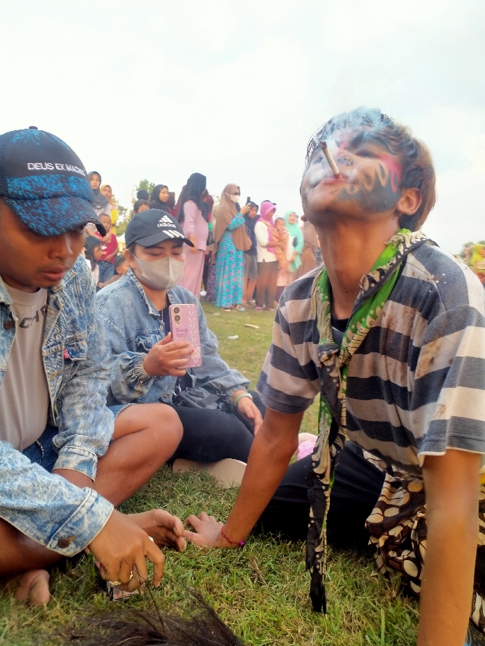 The image depicts an outdoor gathering on a grassy area in the city of Glonggong, Indonesia. In the foreground, three individuals are seated on the green grass. On the left, a male wearing a blue and black "DEUS EX MACHINA" baseball cap and a light-wash denim jacket is manipulating dark, fibrous material on the ground with his hands. In the center, a female wearing a grey face mask, a black Adidas baseball cap, and a light-wash denim jacket sits, holding a pink smartphone with her right hand. On the right, a male with blue and pink face paint on his cheeks and forehead, wearing a grey and white striped t-shirt and a green patterned scarf, exhales smoke upwards from a hand-rolled cigarette held between his lips, with his head tilted back. A dark and light patterned fabric is visible around his lower body. In the background, a crowd of approximately 20-30 individuals, many appearing to be women wearing headscarves (hijabs) and varied colorful clothing, are standing and observing. The sky above is bright and appears overcast. Distant, indistinct vegetation is visible on the horizon.