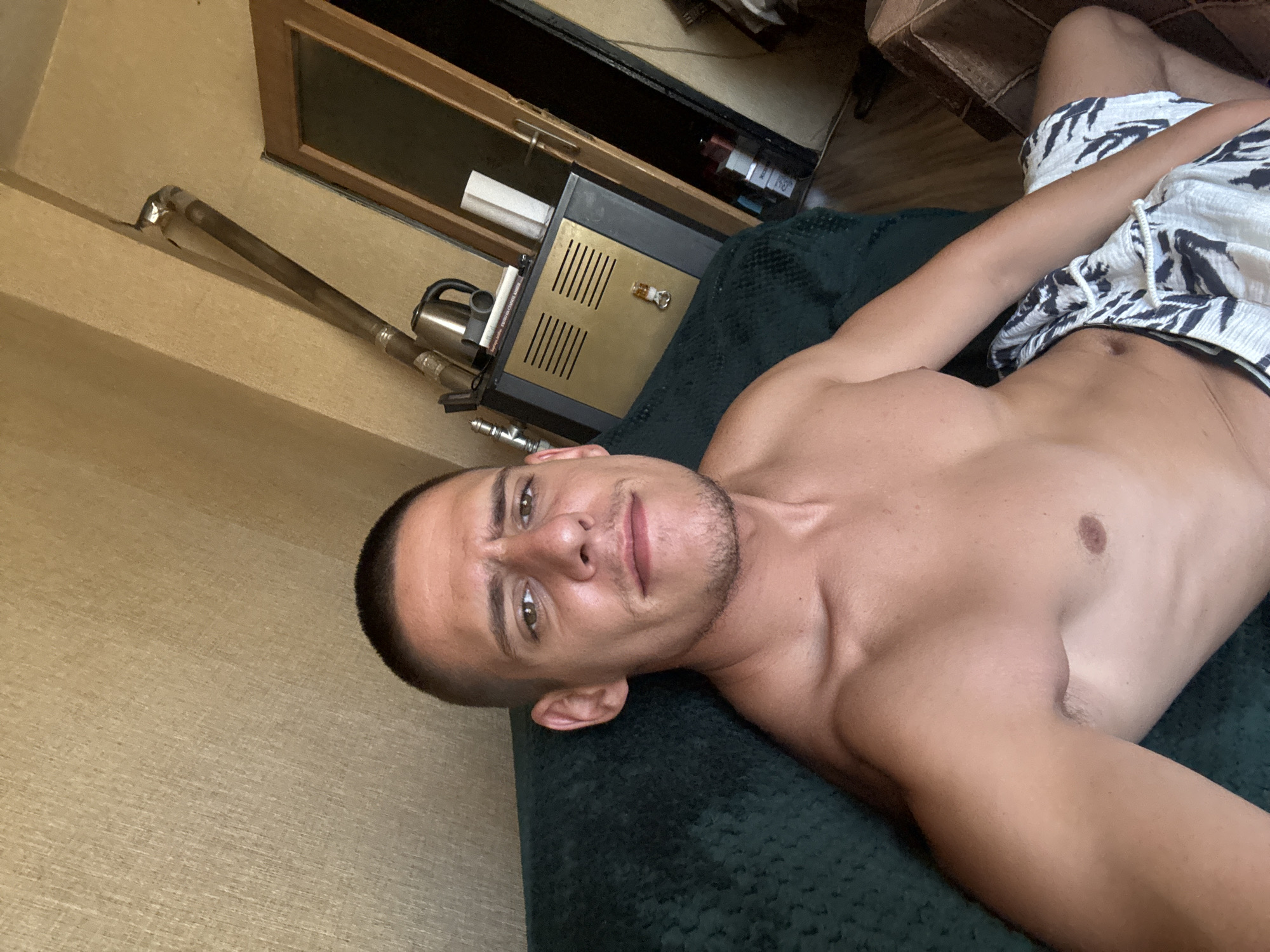 A man is sitting on a dark green sofa. He is shirtless and wearing white and black patterned shorts. He has short, dark hair and is looking towards the camera with a neutral expression. The background shows a textured beige wall, a metal pipe, a black and gold cabinet with a kettle on top, and a wooden door with a glass panel. The scene appears to be an indoor setting.