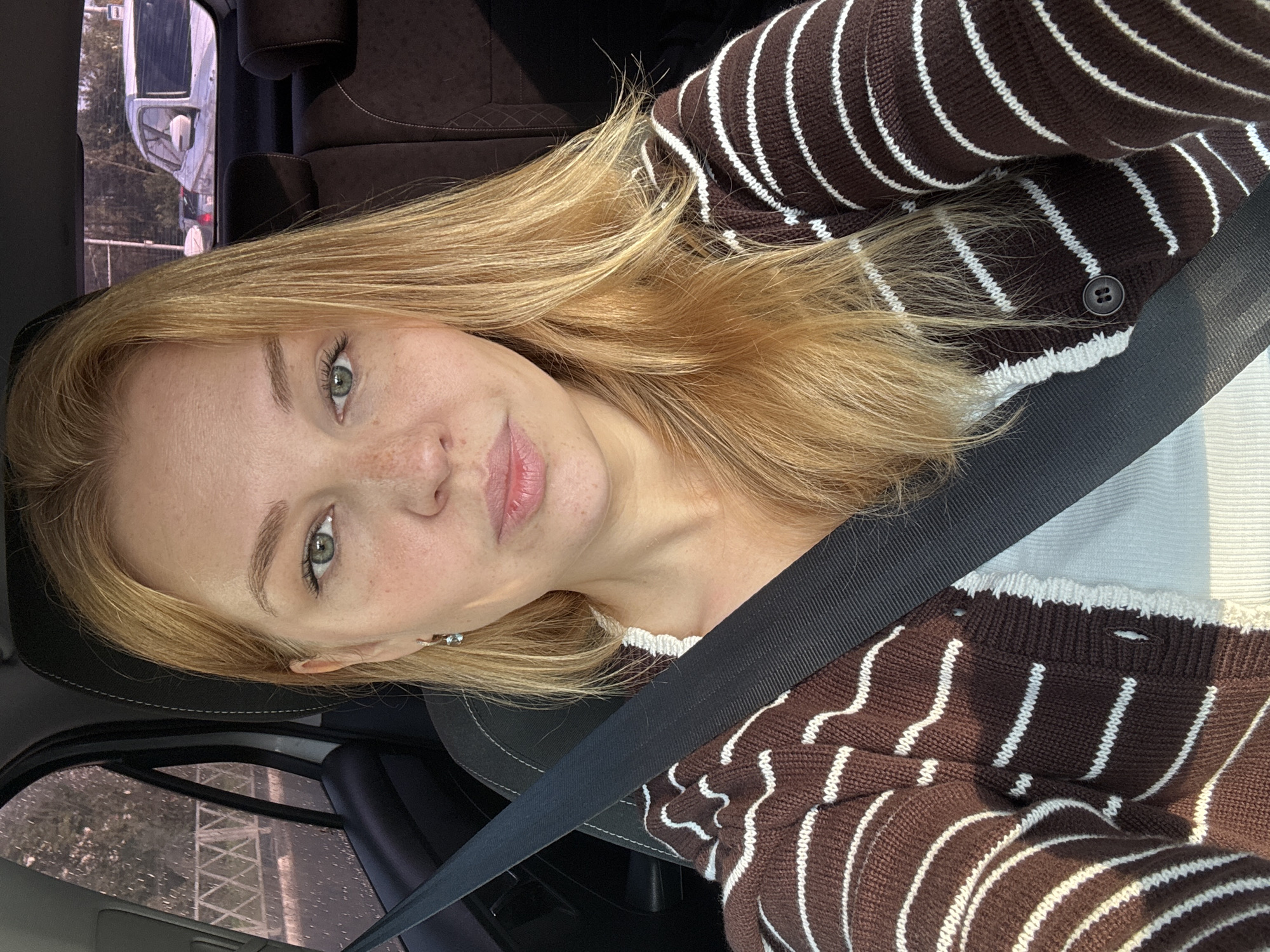 A young Caucasian woman with blonde hair and green eyes is seated in the driver's seat of a car, wearing a brown and white striped cardigan over a white top. She has a black seatbelt across her chest. The interior of the car is visible, including the car's headrests and seats. The background shows a view through the car window of trees and a fence, suggesting an outdoor setting likely within the city of Rusanovo, Russia. The image is a selfie taken by the woman.