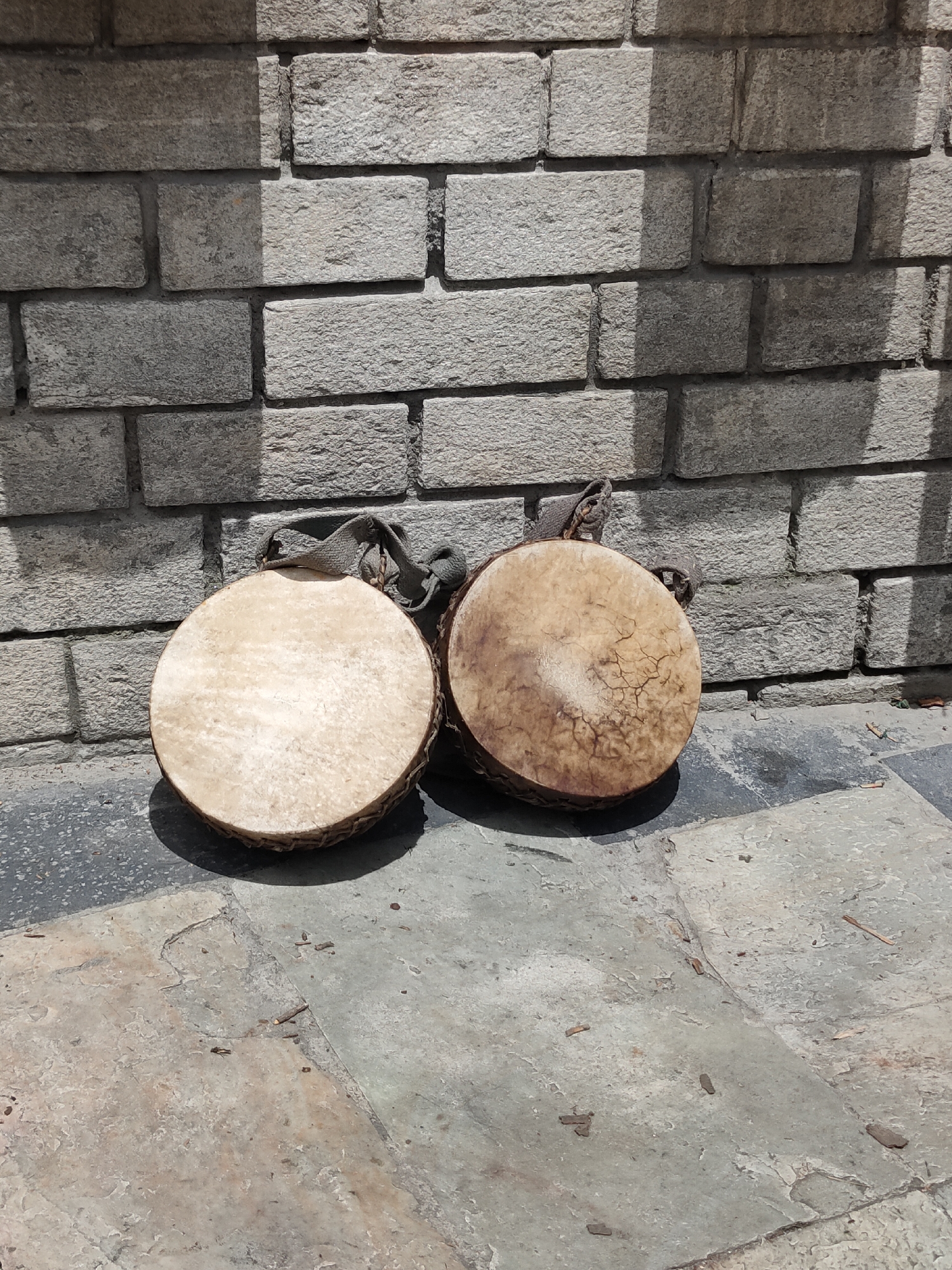 Two small, hand-held frame drums with natural skin heads and woven sides are leaning against a rough-hewn stone brick wall. One drum is lighter in color with a smooth, slightly worn surface, while the other is darker and shows a network of cracks. Both drums have simple fabric straps attached to them, suggesting they are meant to be carried or hung. The drums are resting on a paved ground surface made of large, irregular stone tiles. The scene is illuminated by strong sunlight, casting distinct shadows from the drums and a vertical element on the right side of the frame onto the wall and ground. The background consists of the textured stone wall, with visible mortar lines between the rectangular bricks. The ground is somewhat uneven, with small debris scattered on its surface. The overall impression is one of an outdoor setting, possibly in a historical or cultural area, given the style of the wall and the instruments.