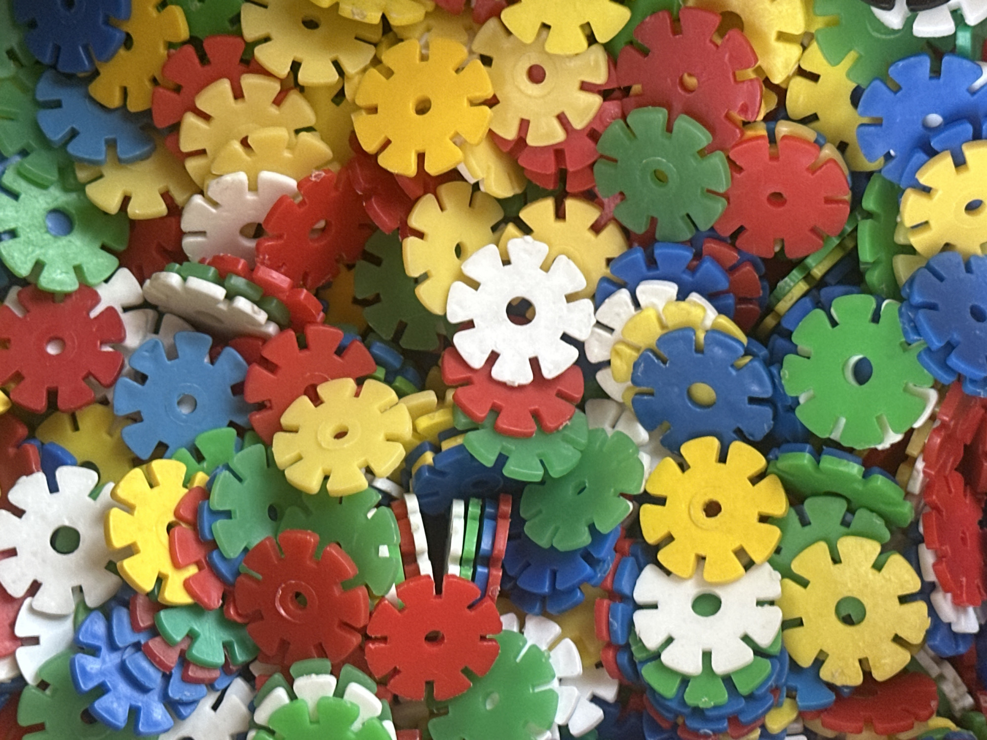 A close-up, overhead shot displays a dense pile of colorful, plastic snowflake-shaped construction toys. The toys are predominantly red, yellow, green, and blue, with a few white ones visible. They are stacked and intertwined, creating a textured and vibrant mosaic. The lighting is bright, casting subtle shadows on the edges of the interlocking pieces. No people, city, or environmental context are discernible in this image.