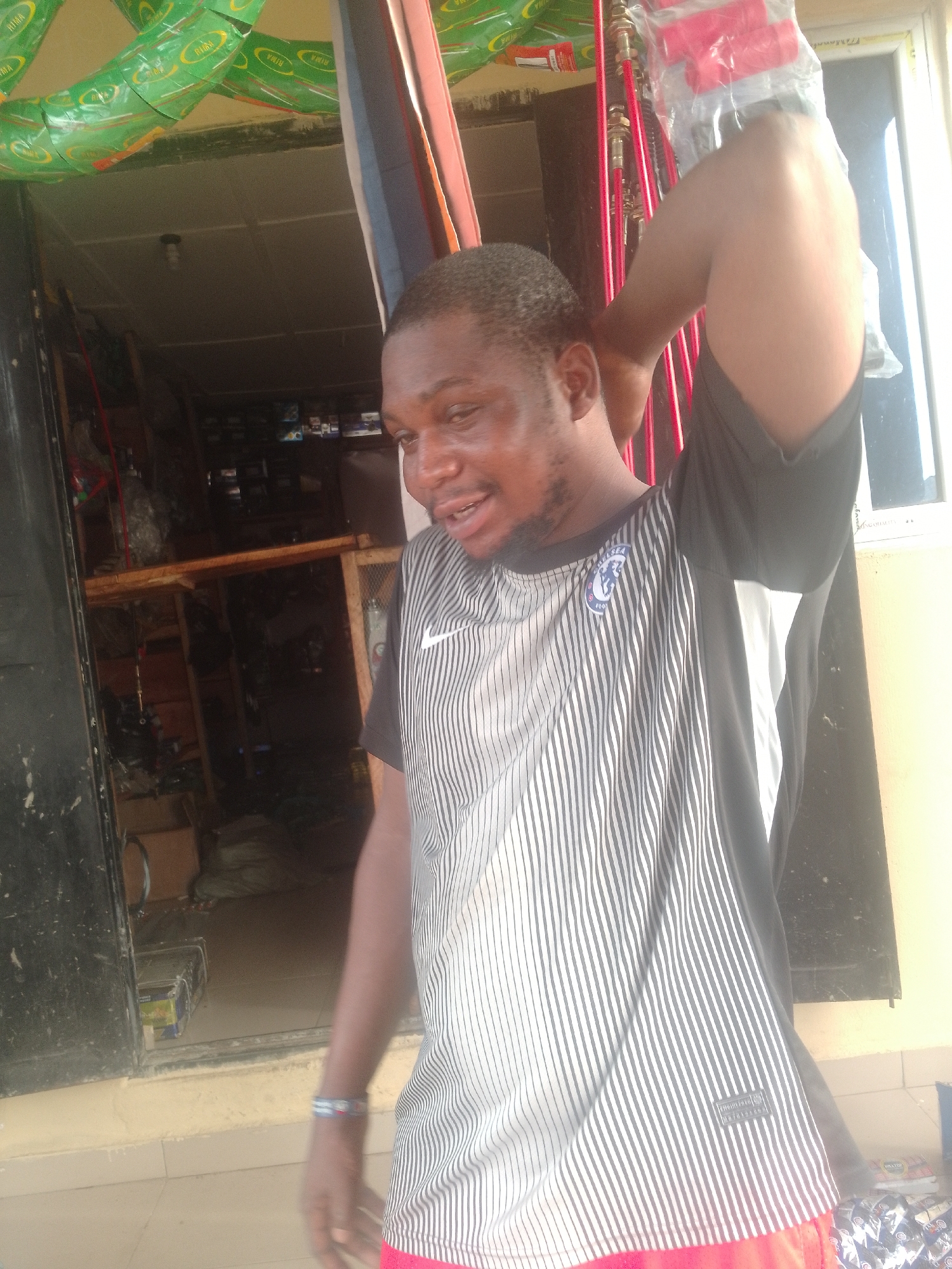 A man stands near the entrance of a shop in Adankolo, Nigeria. He wears a black and white striped shirt with a Chelsea logo and red shorts. His right arm is raised, holding a plastic-wrapped item. The shop behind him contains various plumbing supplies and other goods. The entrance is decorated with green and yellow packaging.