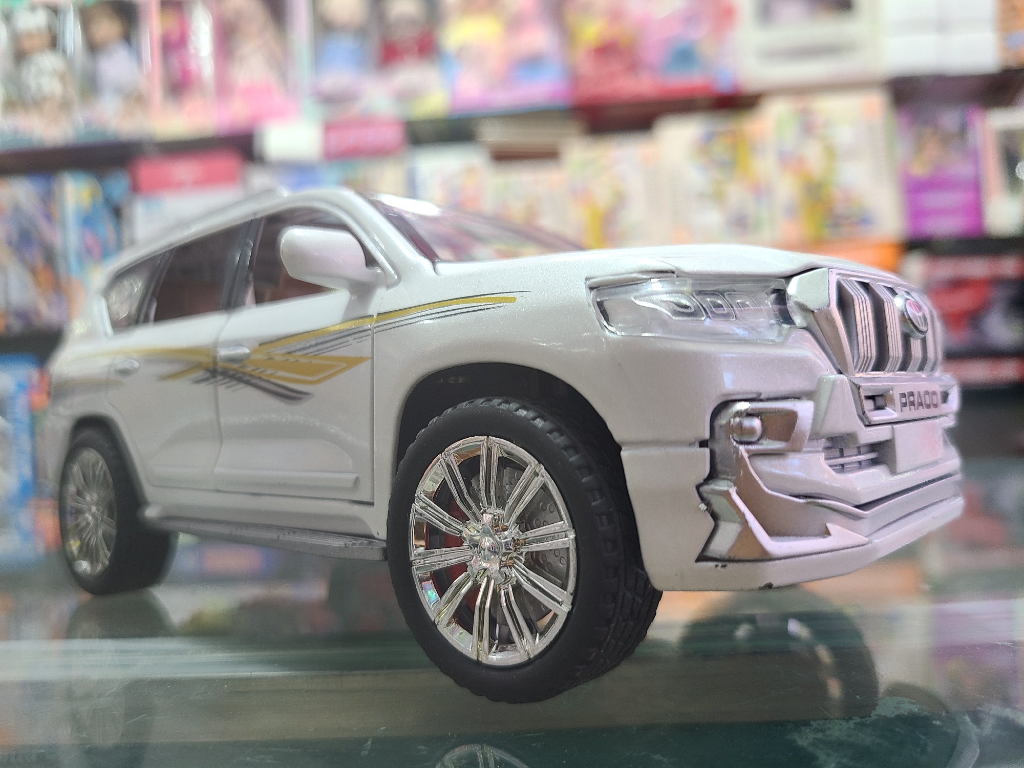 A close-up shot features a white toy car, specifically a model of a Toyota Land Cruiser Prado, with decorative gold and black stripes on its side. The car is positioned on a reflective surface, possibly glass or a similar material. In the blurry background, a shelf is visible, filled with books and various boxed items, typical of a store environment. The location is presumed to be a shop in Quetta City Tehsil, Pakistan.