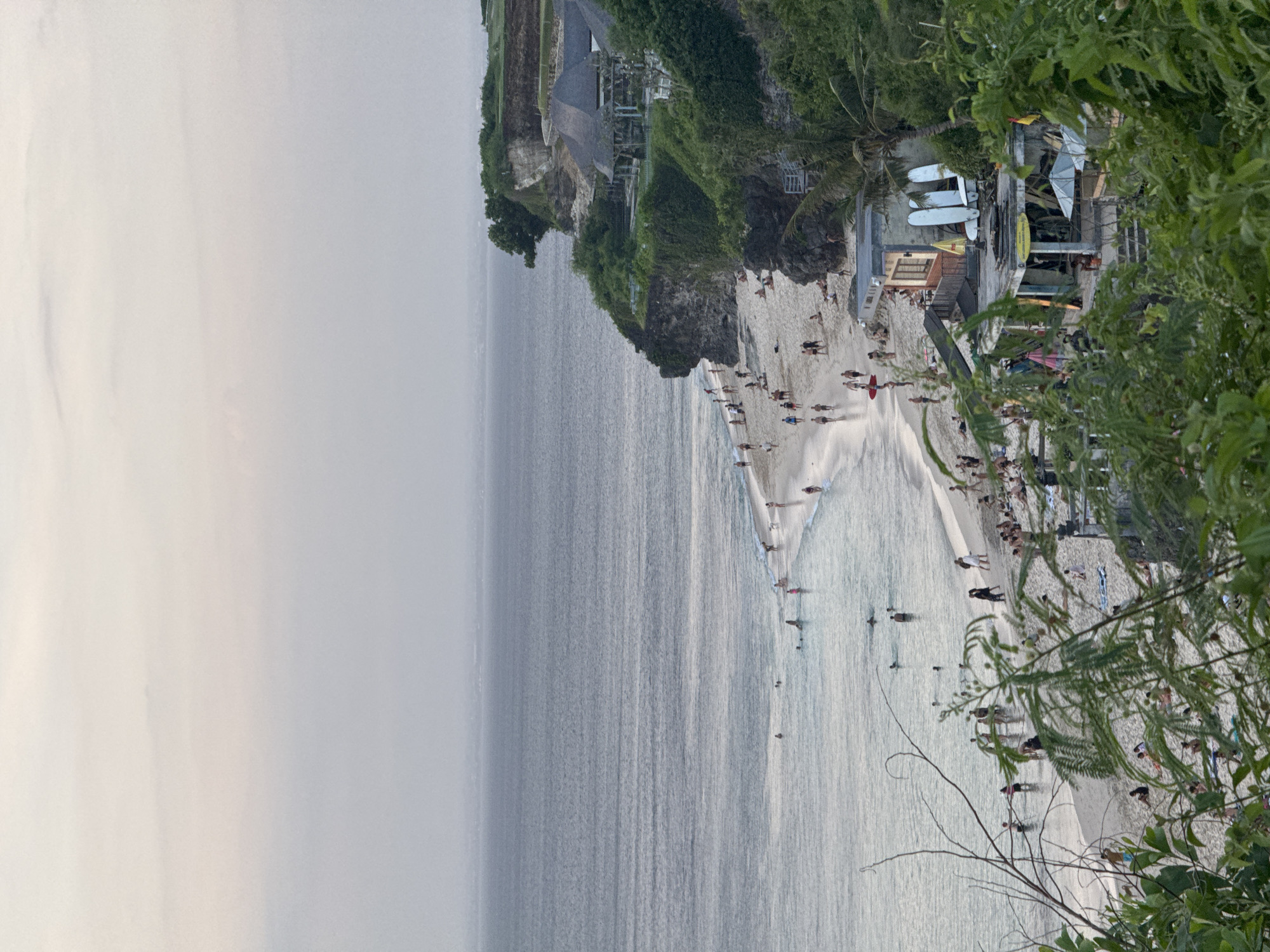 The image shows a view of Uluwatu Beach in Kuta Selatan, Indonesia from an elevated position. The sky is overcast. The ocean is a gray-blue color with slight waves. A sandy beach stretches along the coast. There are many people on the beach, some swimming, some walking, and others sunbathing. In the upper-right portion of the image, there are green plants and trees. There is a structure that appears to be a building above the beach on the cliffside, with surfboards on top. The scene has a cloudy, slightly hazy atmosphere.
