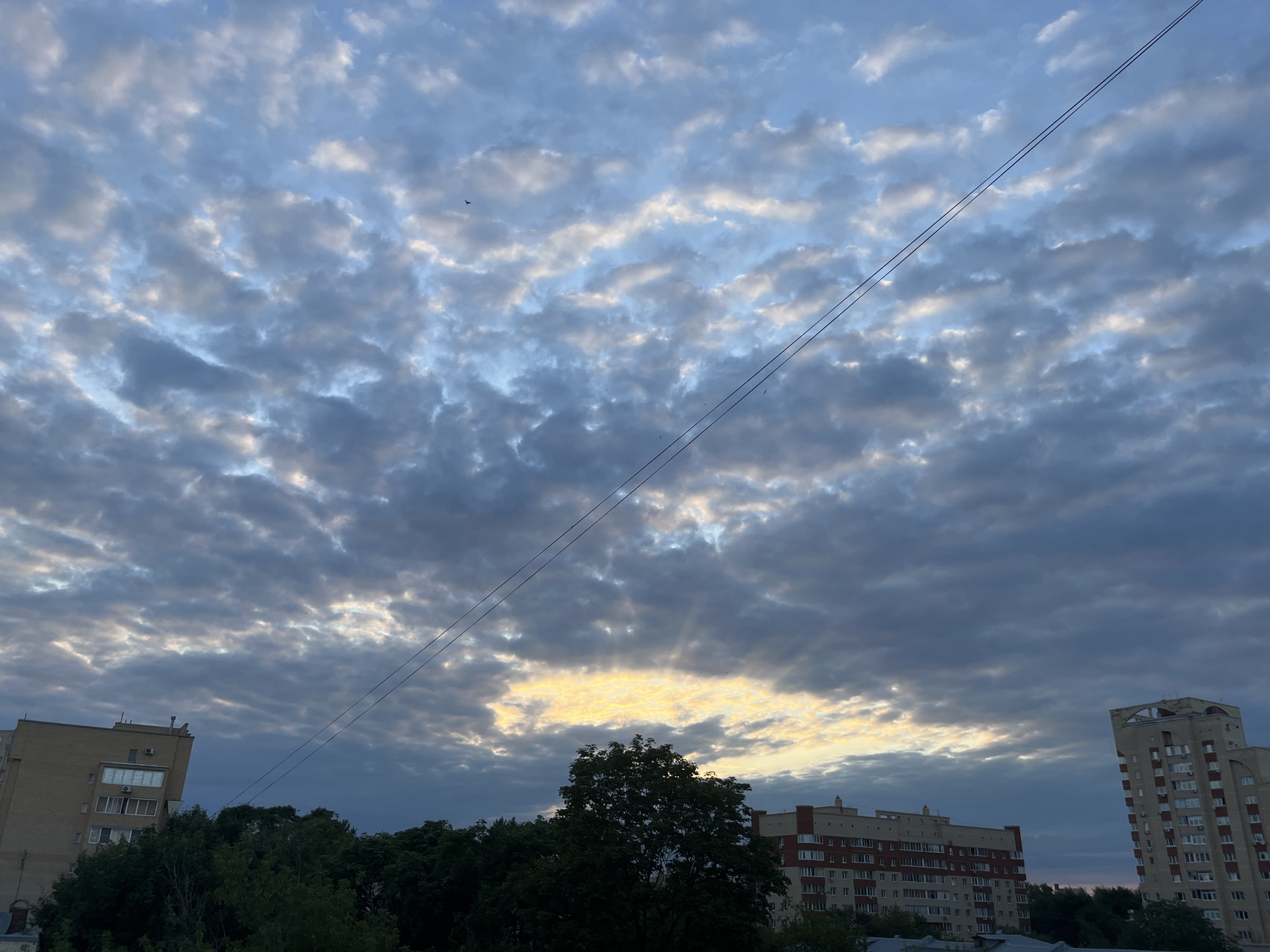 The image depicts an overcast sky with visible sunbeams breaking through a gap in the clouds, suggesting it might be dawn or dusk. Two multi-story residential buildings, characteristic of Soviet-era architecture, are visible in the foreground, one on the left and another on the right. A dense canopy of green trees separates the buildings from the sky. Power lines stretch diagonally across the frame. The scene is likely located in Ivanteyevka, Russia, given the architectural style and urban setting.