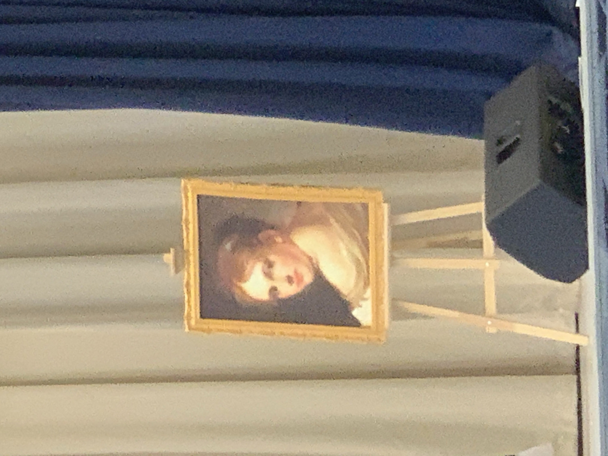 The image displays a small, framed painting mounted on a wooden easel, set indoors. The painting, presented in an ornate golden frame, features a head-and-shoulders portrait of a young person, likely a child or cherub. This individual has light hair and fair skin, with soft features, and appears to be gazing upwards and slightly to the viewer's right, reminiscent of a classical or Baroque artistic style.
Behind the painting, a light-colored, possibly white or beige, pleated fabric or curtain forms the background. Above this, a darker blue, similarly pleated fabric or curtain spans across the upper portion of the scene. On the right side of the image, partially visible, is a dark grey or black rectangular speaker or piece of sound equipment, suggesting the setting might be an event or presentation space where audio amplification is used.
The overall lighting appears dim, contributing to a slightly grainy quality in the photograph. The scene suggests an art display or exhibition within a formal or semi-formal venue. There is no visible text in the image. The location is contextualized as Chaikovsky, Russia.