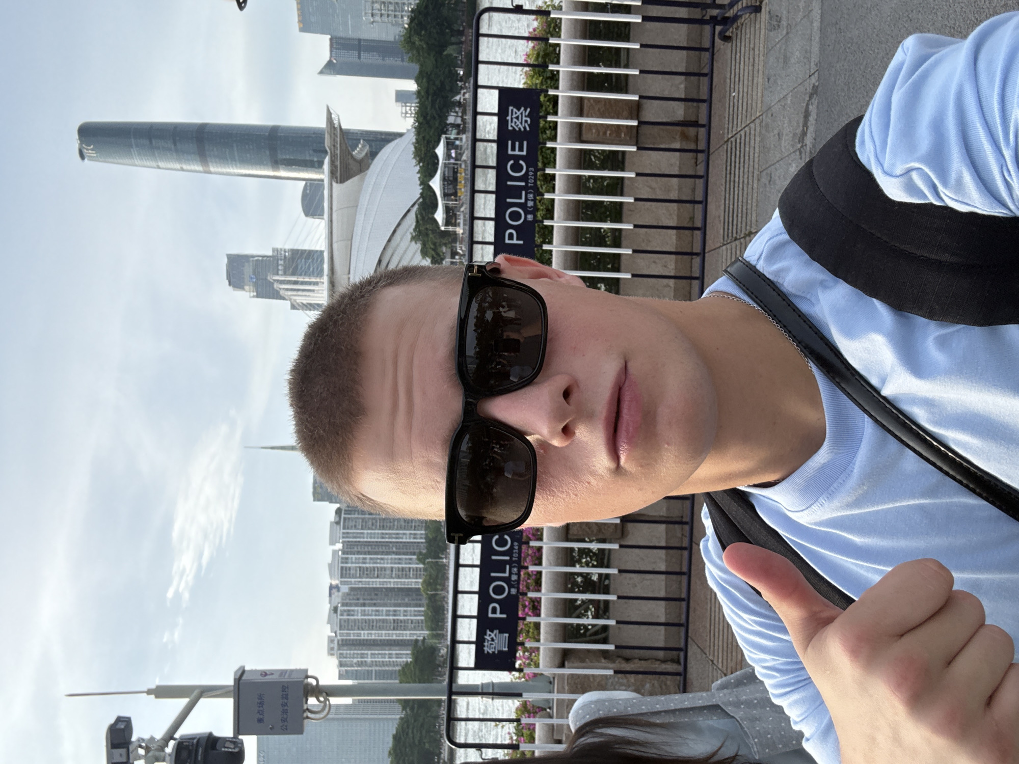 A young man with short brown hair and sunglasses is taking a selfie. He is wearing a light blue t-shirt and a black backpack. He is giving a thumbs up with his left hand. In the background, there is a fence with a sign that says "POLICE" in English and Chinese. Behind the fence, there is a river and a cityscape with tall buildings. The man is in Haizhu District, China. The scene appears to be a tourist area with a police presence.