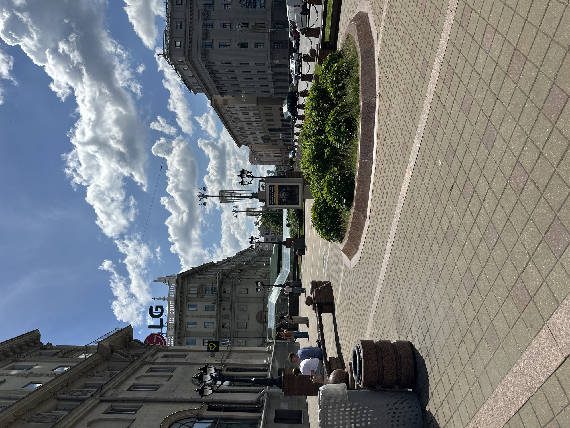The image captures a street scene in Minsk, Belarus, under a bright, partly cloudy sky. The perspective is rotated, giving a vertical orientation to the buildings and street. The foreground is dominated by a tiled pedestrian walkway with a decorative border. A large, circular planter with lush green bushes is set into the walkway.
To the right of the planter, a peculiar object resembling stacked tires or weights is visible, with a few people gathered around it, seemingly engaged in an activity that might involve exercise or demonstration. One person in a white shirt and another in a blue shirt are bent over the object. Further down the street, more people are seen walking and standing.
The background features grand, multi-story buildings with ornate architecture, characteristic of a city center. One building prominently displays an "LG" logo and a red circular logo, indicating a commercial presence. Streetlights and a billboard add to the urban landscape. The sunlight suggests it is daytime, likely midday given the brightness and shadows. The presence of clouds indicates dynamic weather conditions. The overall atmosphere appears to be casual and active, with people enjoying a pleasant day outdoors in a city environment.